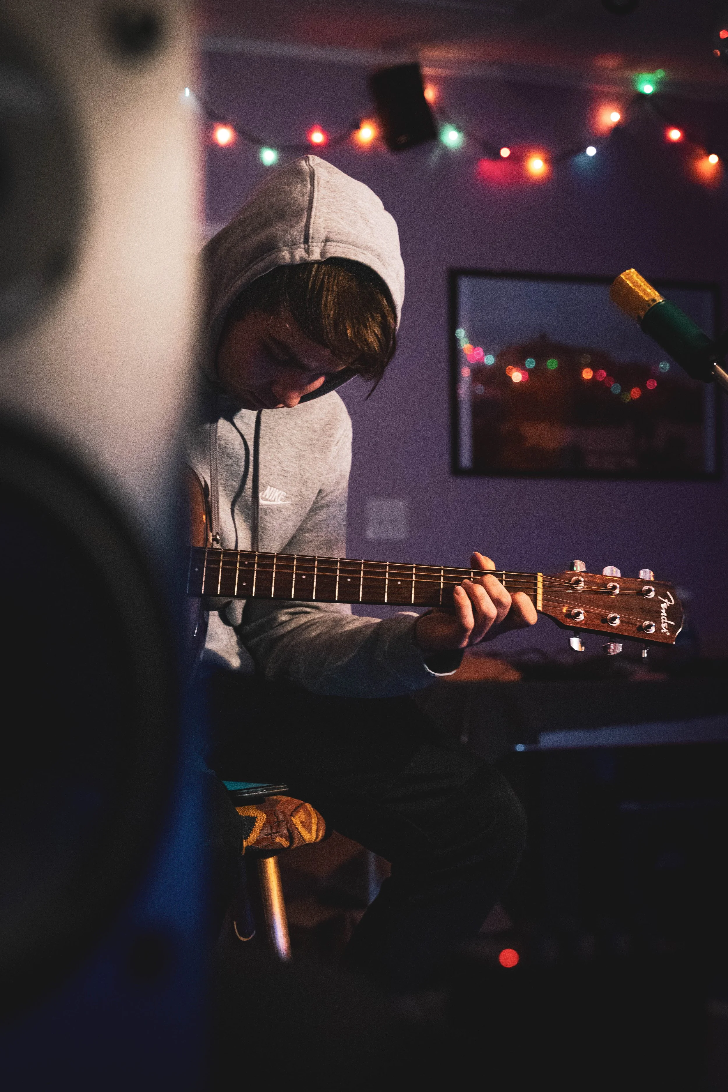 A young man wearing a gray hoodie playing an acoustic guitar in a dimly lit room decorated with colorful string lights.