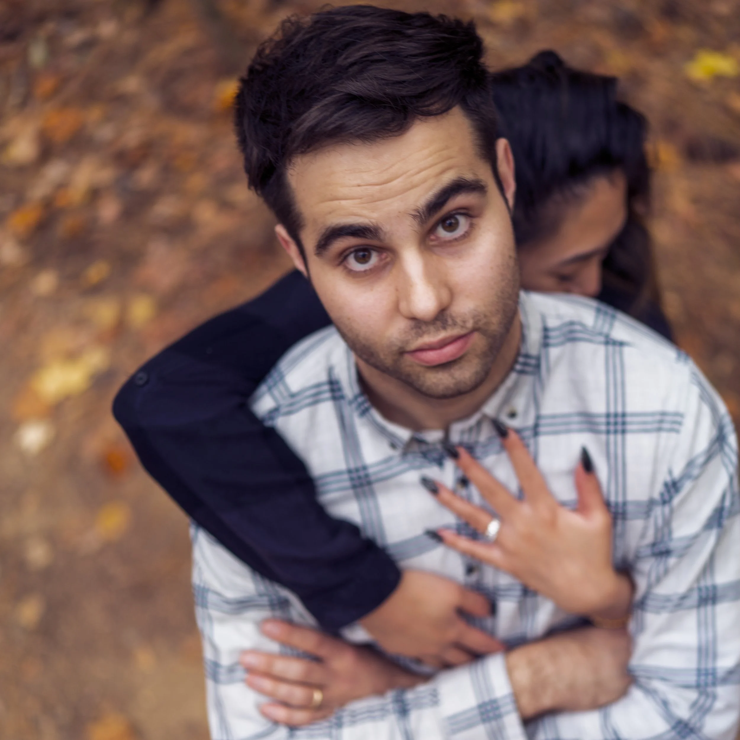 A young man with dark hair and light skin looking up into the camera, with a woman embracing him from behind with her hands on his chest, outdoors with autumn leaves on the ground.