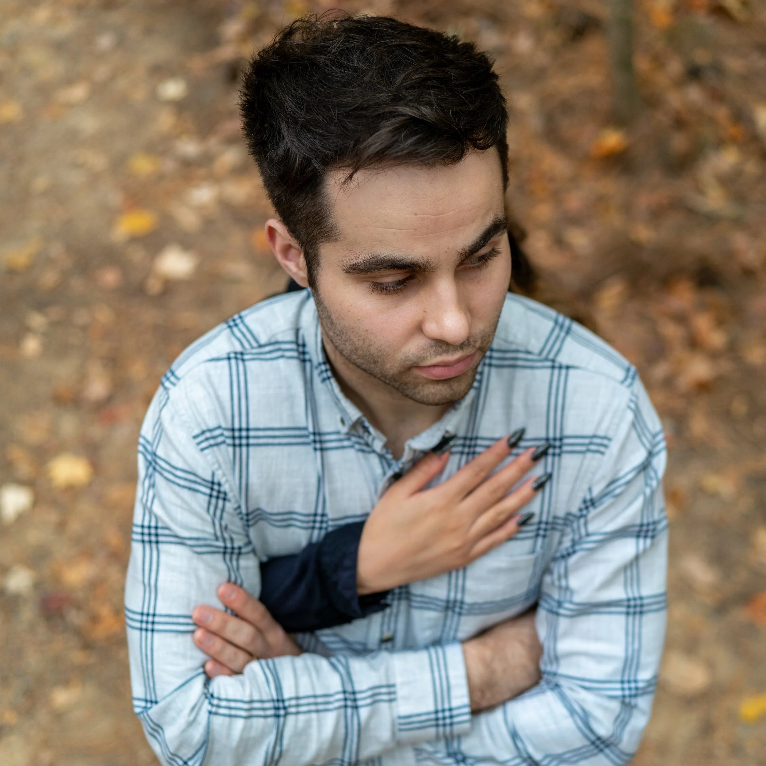 A young man standing outdoors on a fall day, wearing a plaid shirt, with one hand on his chest and the other arm crossed over his chest.