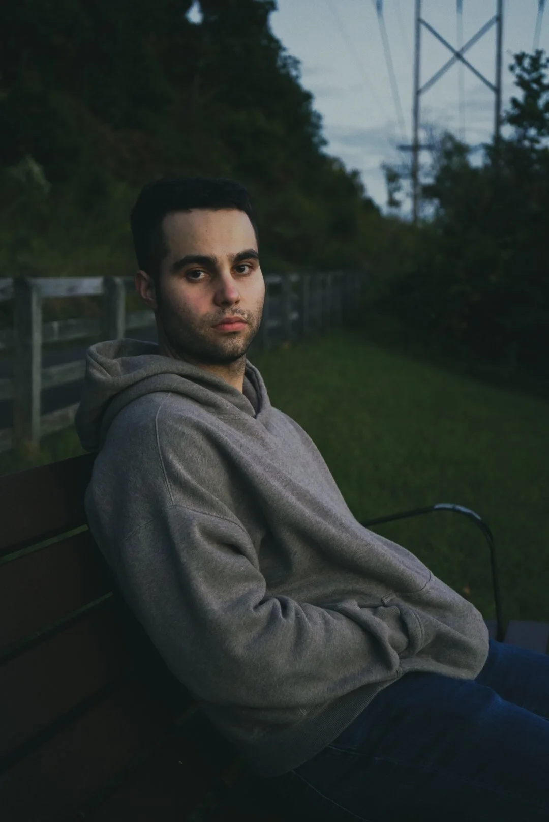 A young man with dark hair and light skin sitting on a park bench during dusk, wearing a gray hoodie and jeans, with a grassy area, a wooden fence, and power lines in the background.