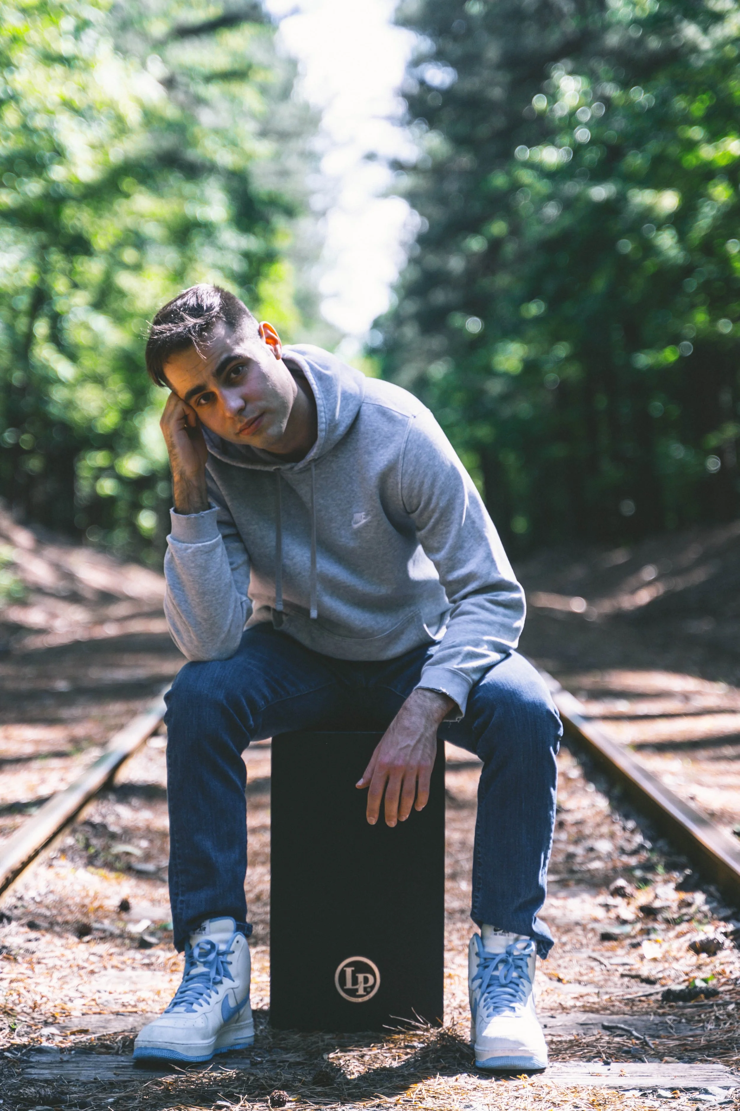 A young man in a gray hoodie and jeans sitting on a black box on a railroad track in a forest, with trees and greenery in the background.