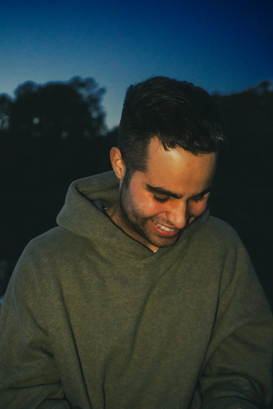A man with dark hair and a beard wearing a gray hoodie, looking down with a slight smile, against a dark outdoor background with trees and a blue sky.
