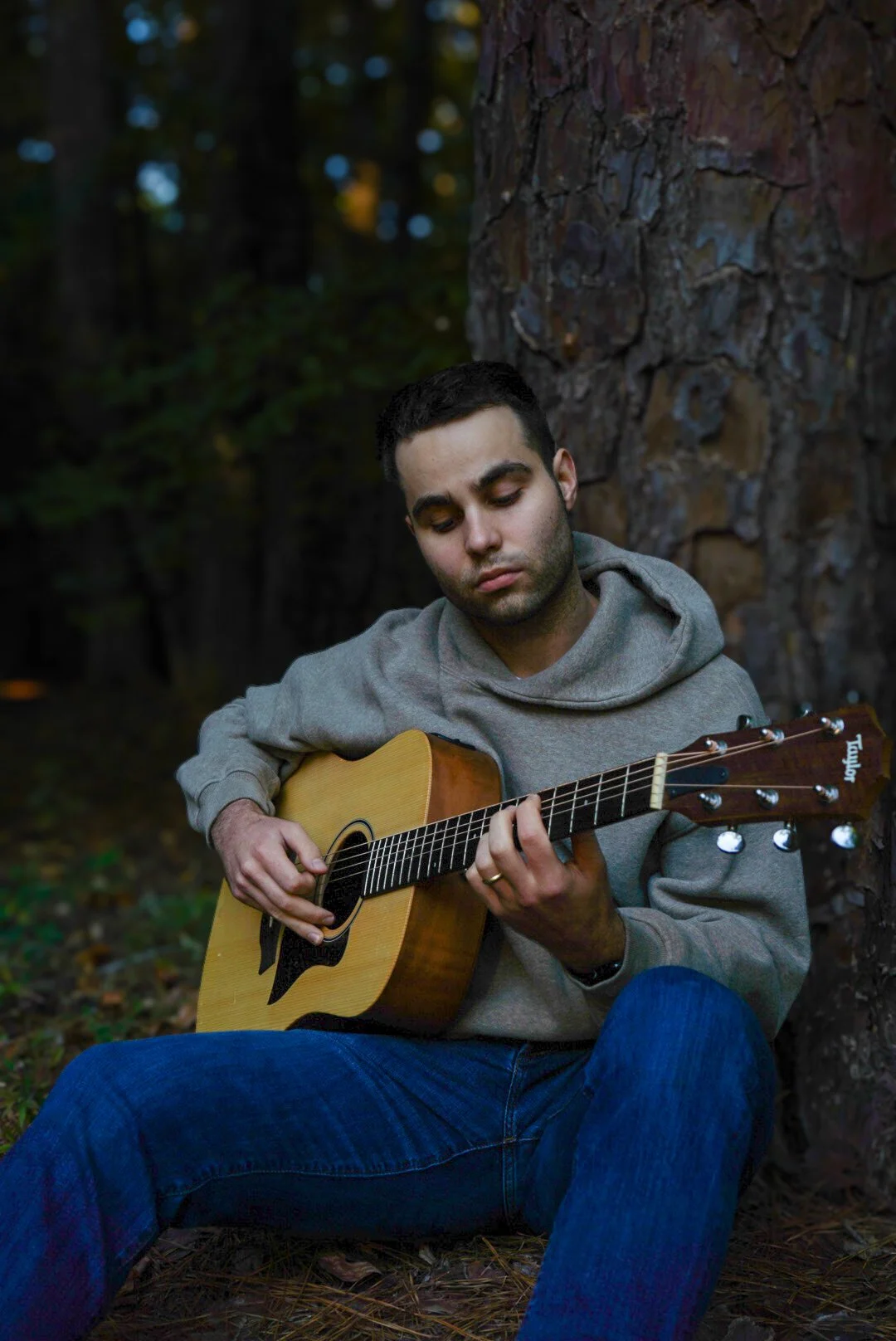 A young man sits outdoors in a forest, playing an acoustic guitar next to a tree, with a focused expression.