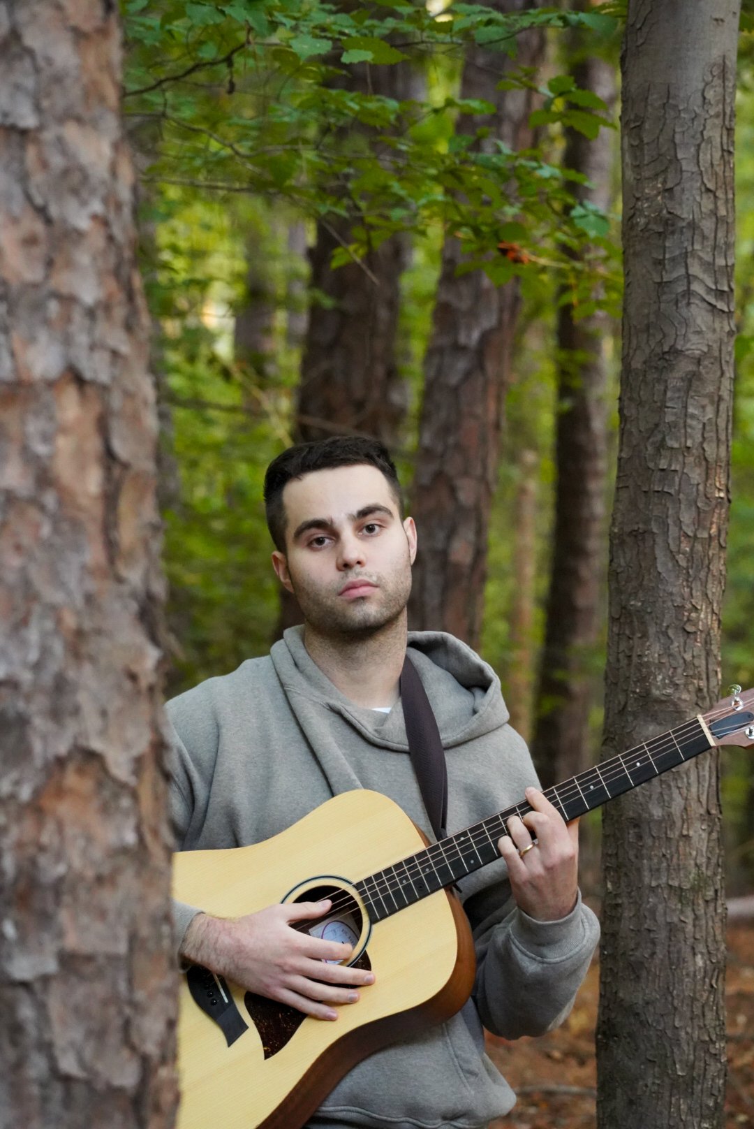A young man playing an acoustic guitar in a forested area surrounded by trees.