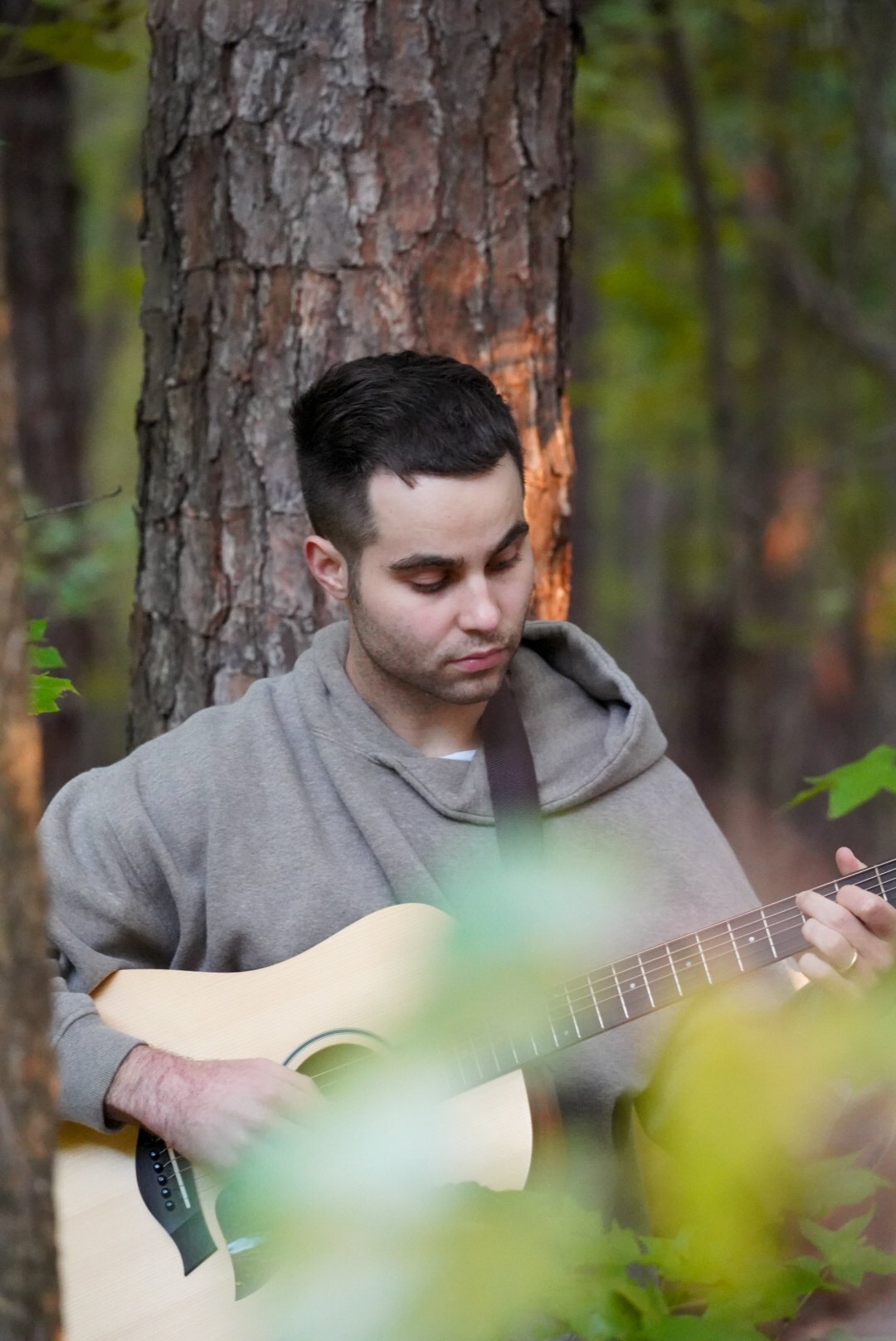 A young man with short dark hair and light skin playing an acoustic guitar in a forest, standing against a large tree, wearing a grey hoodie, with green foliage in the foreground and background.