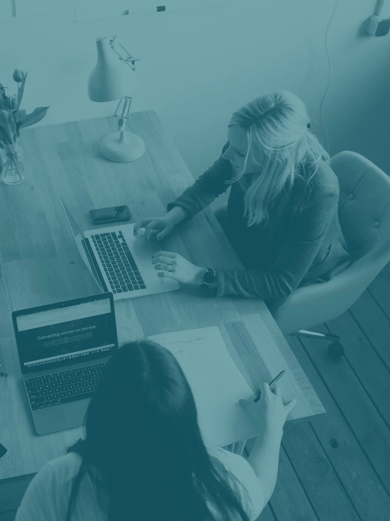 Two women sitting at a wooden table working on laptops, with one woman using a smartphone and a notebook, in an office setting.