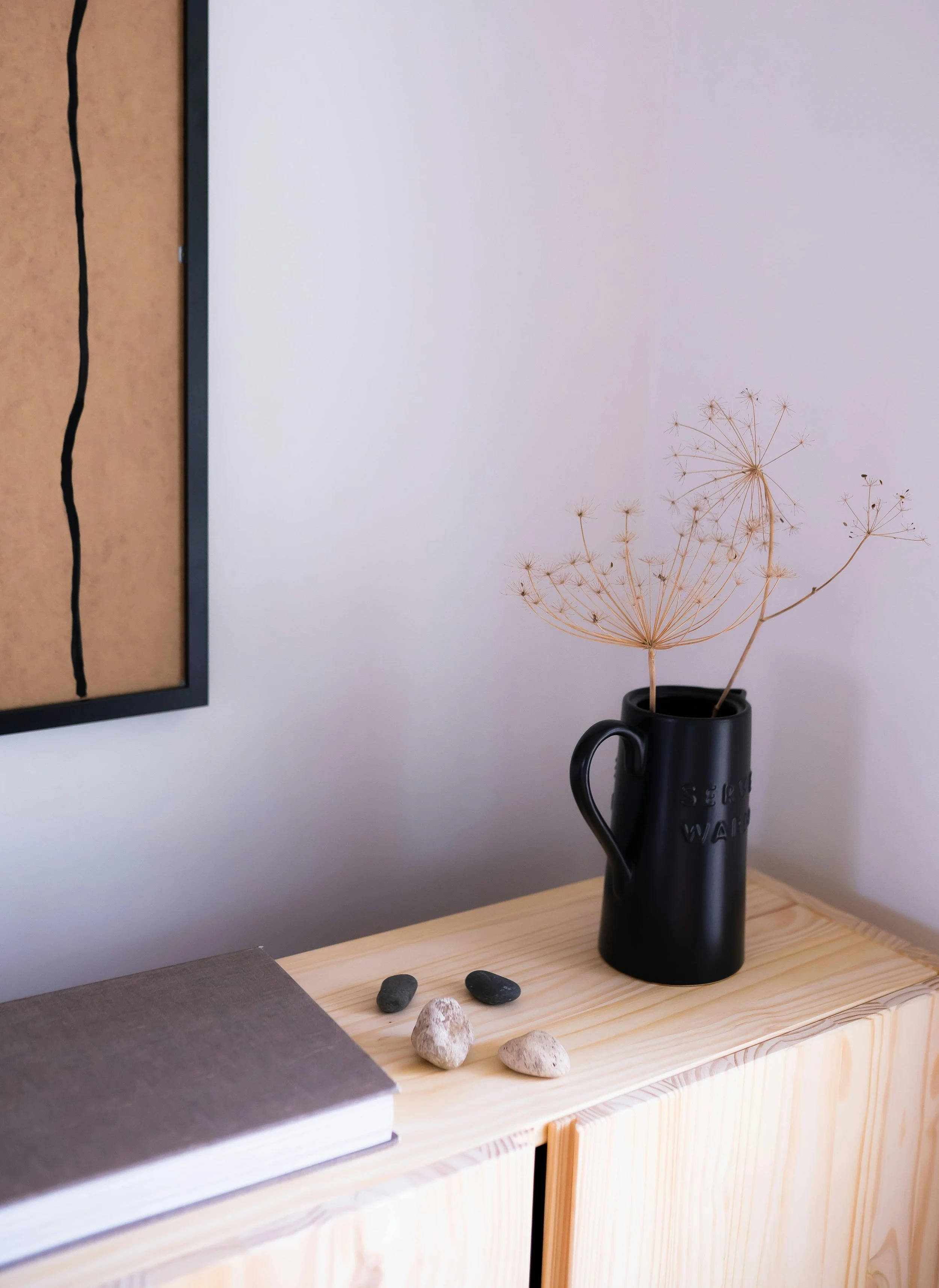 A black vase with dried flower stems on a wooden surface, beside a closed book and several small rocks. Part of a framed art piece is visible on the wall.