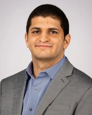 A man with short dark hair wearing a light blue dress shirt and a gray suit jacket, smiling against a light gray background.
