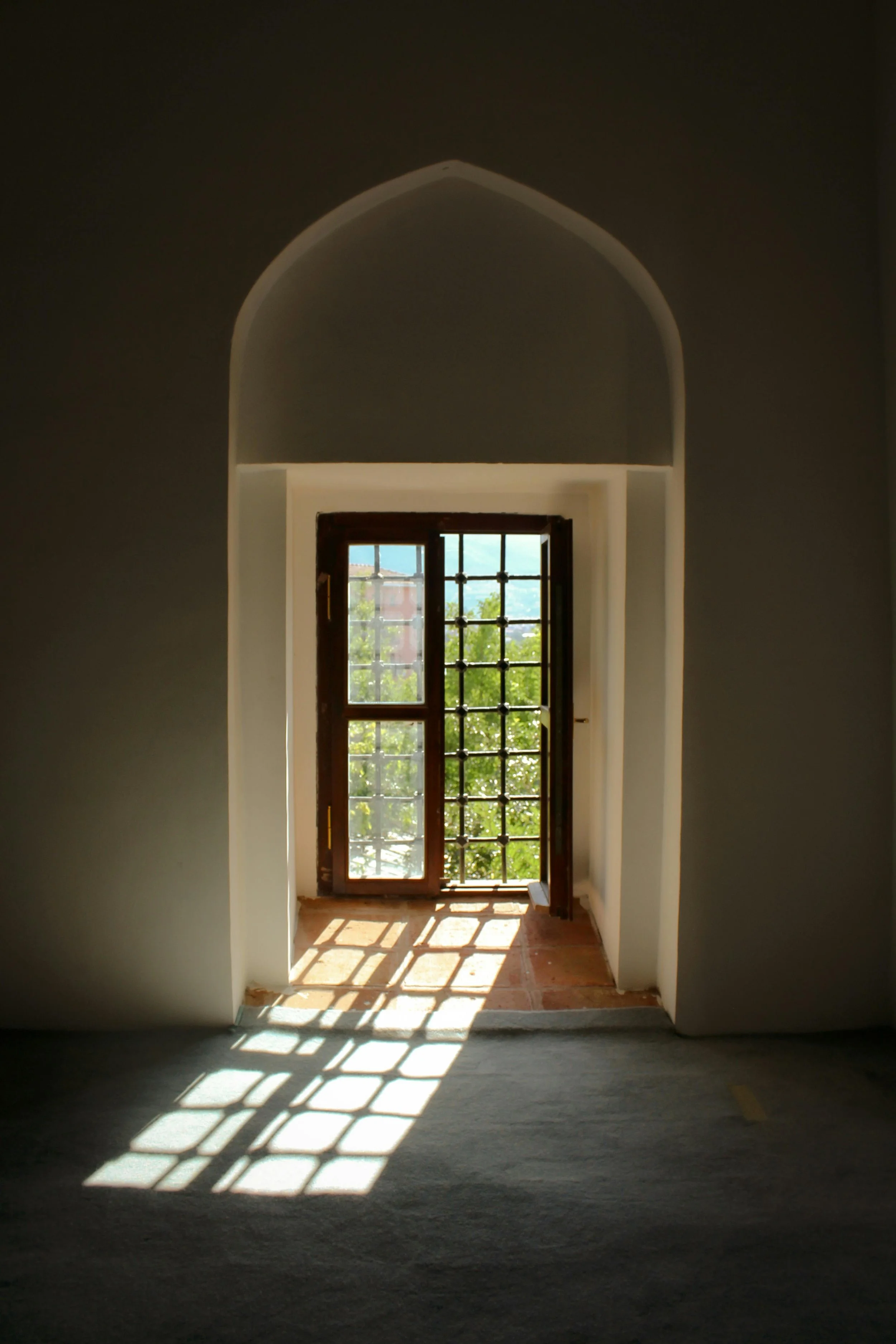 Interior view of a room with a large arched window, sunlight casting a patterned shadow on the floor, and greenery visible outside.