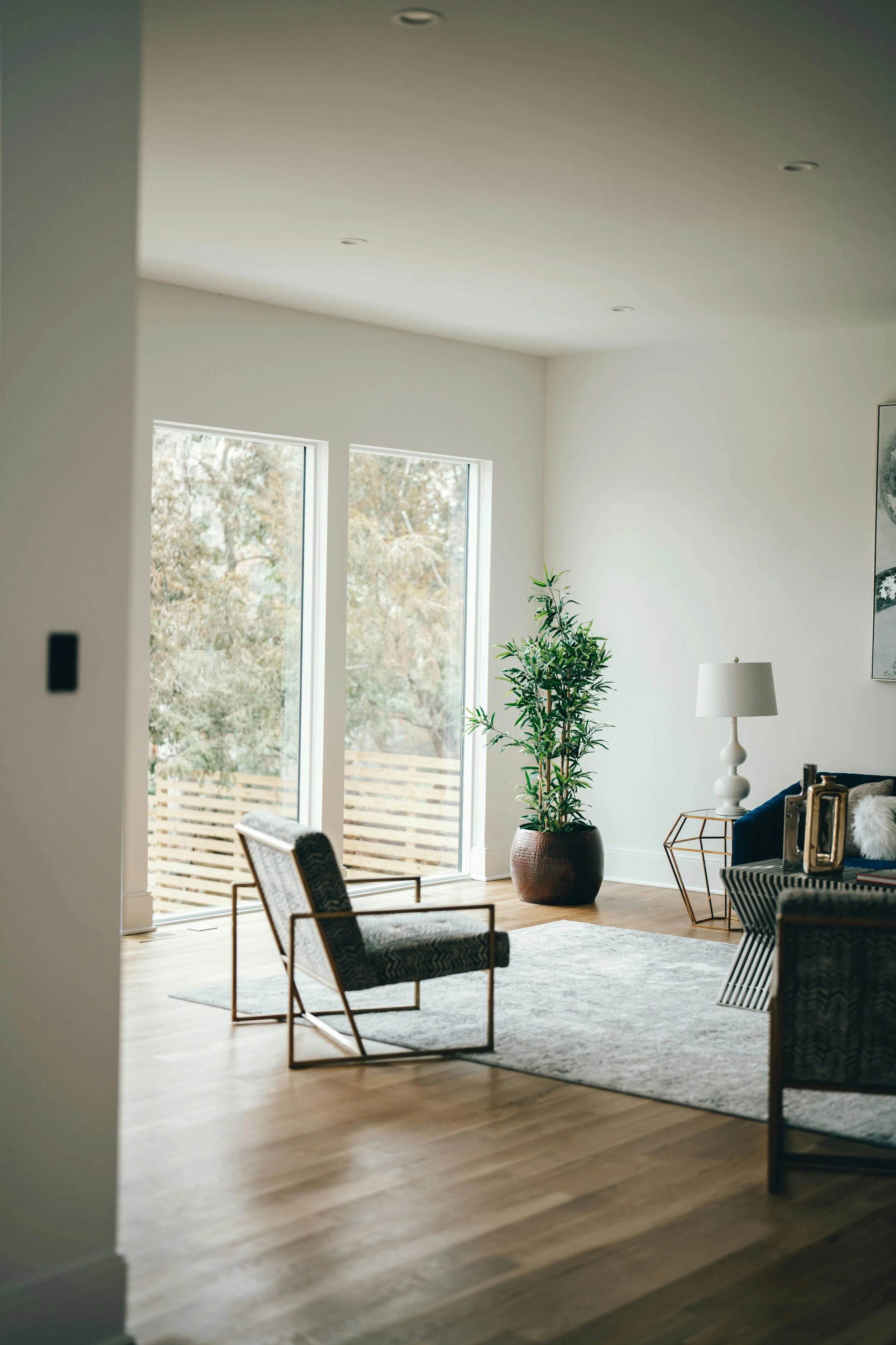 Modern living room with wooden floors, large sliding glass doors, a potted plant, a white table lamp, and seating furniture.