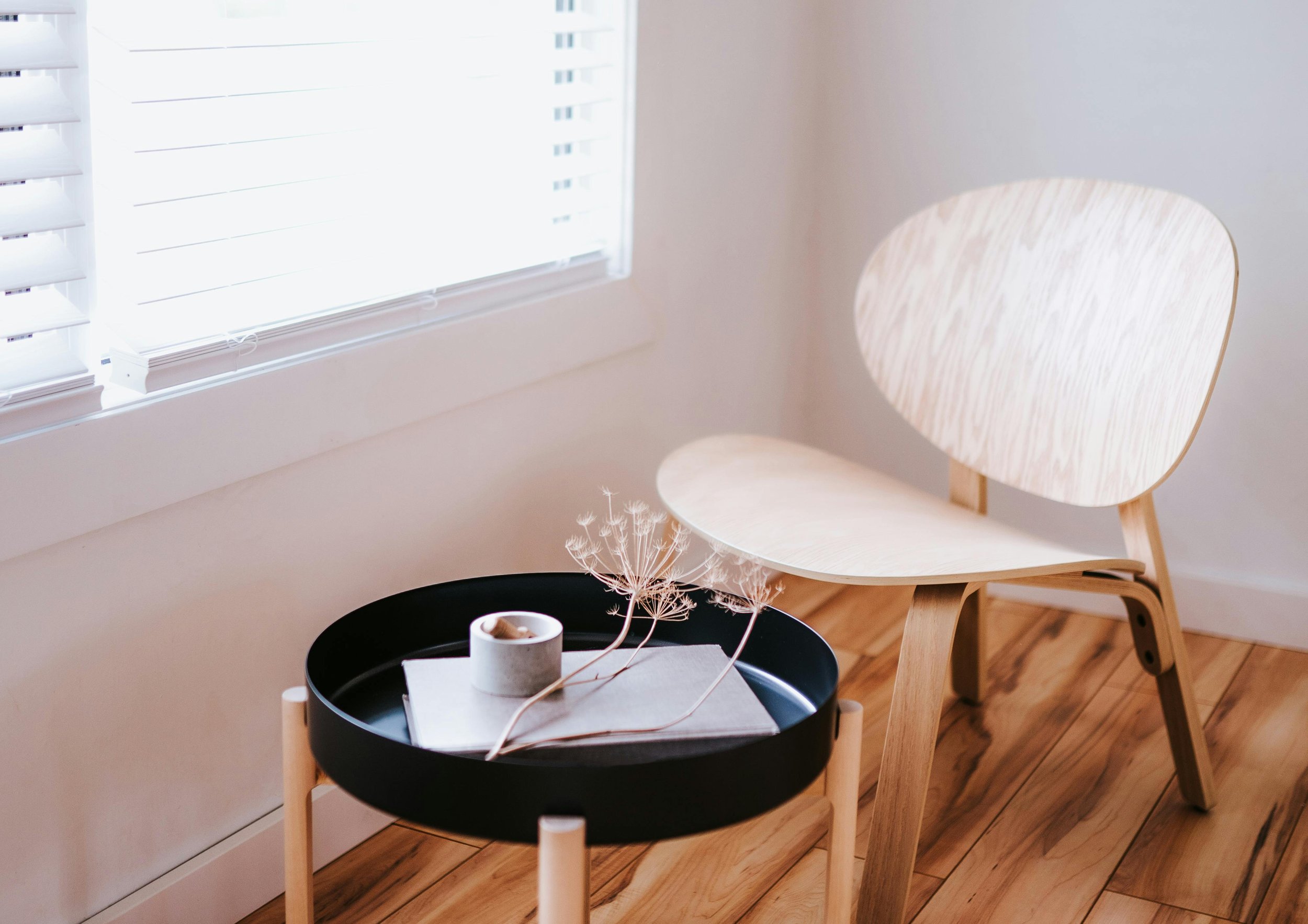 A minimalist interior with a light wooden chair next to a black side table holding a small plant, a rolled paper, and a book, near a window with white blinds.