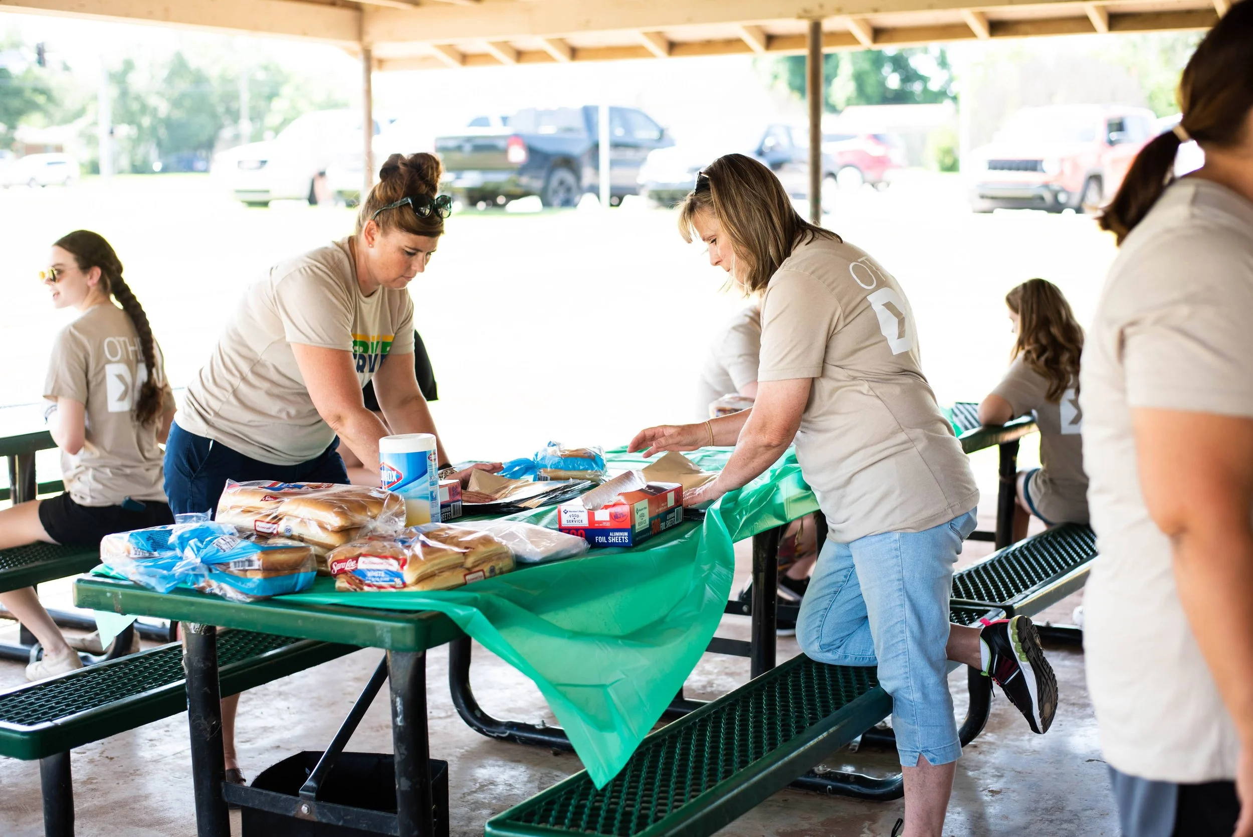 People preparing food at an outdoor covered picnic area, with three women standing and handling supplies on a green plastic table, surrounded by benches and parked cars in the background.