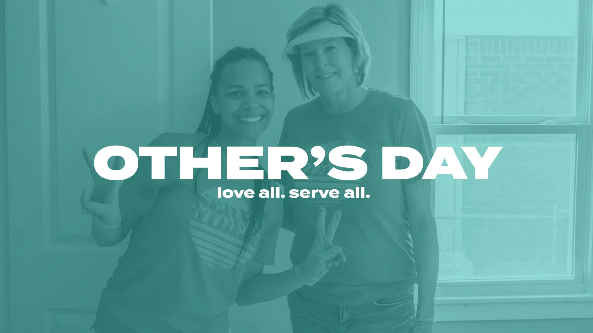 Two women smiling and making peace signs, wearing gloves, with a background of a window and brick wall, promoting Others' Day with the slogan "love all. serve all."