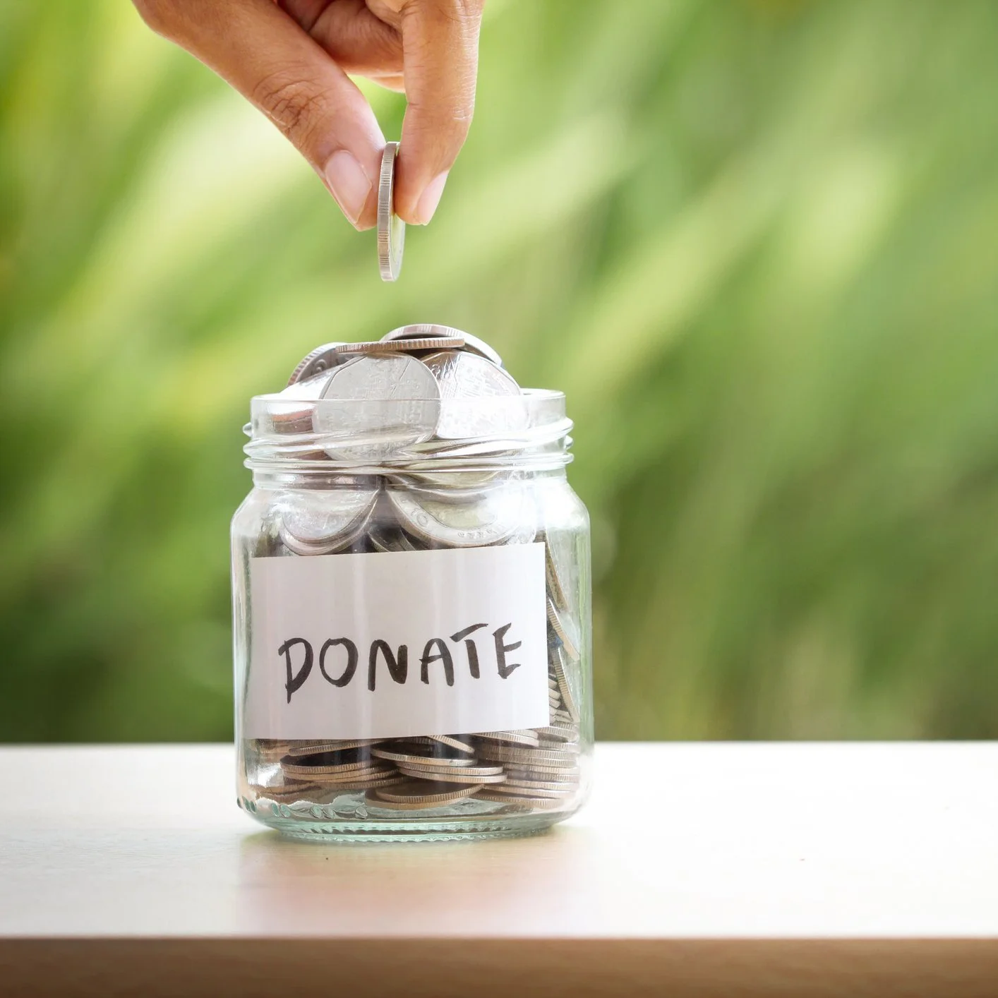 Hand dropping a coin into a glass jar labeled 'DONATE', filled with coins, on a white surface with a blurred green background.