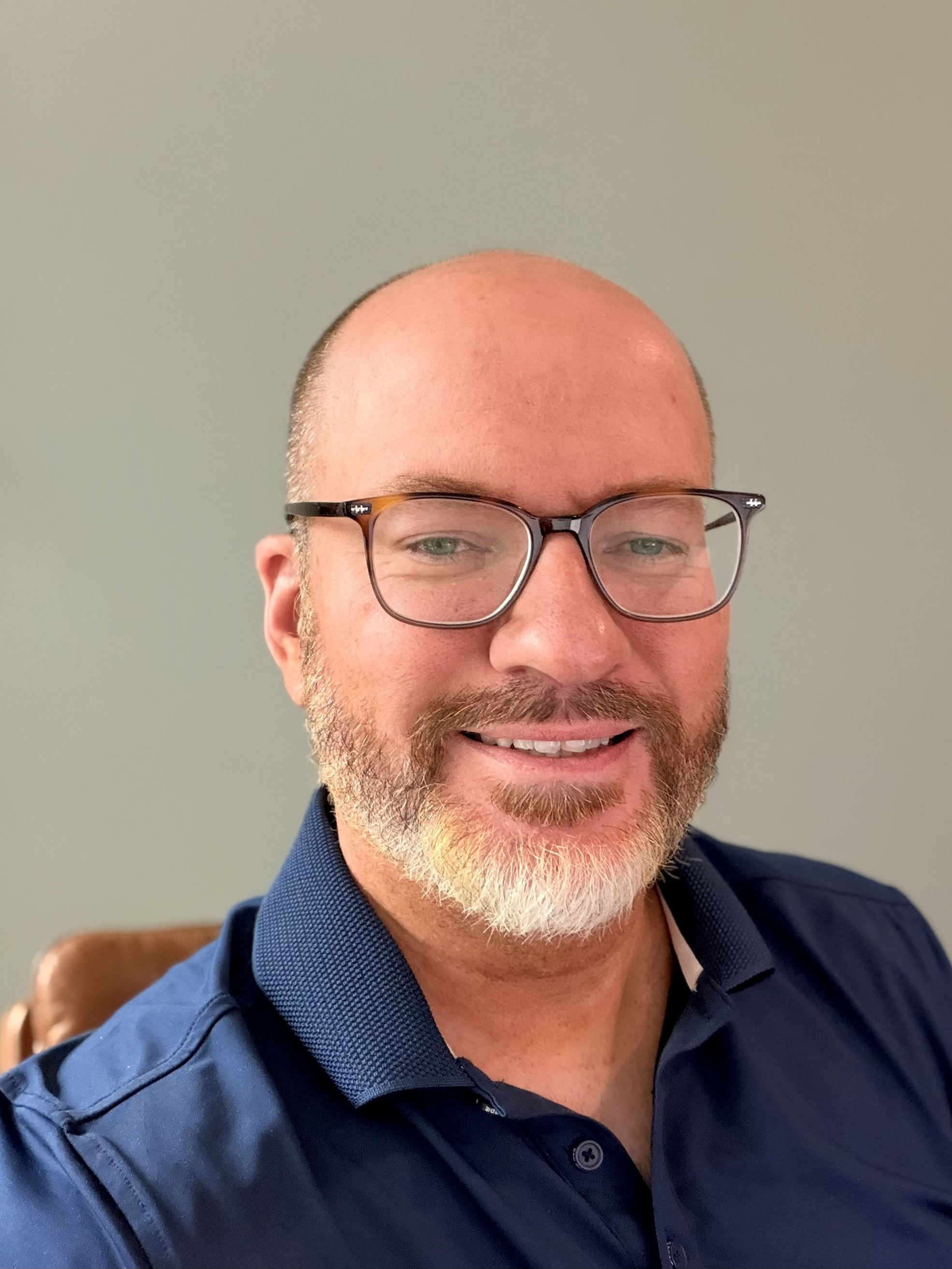 Close-up photo of a smiling man wearing glasses, with a beard and mustache, dressed in a navy blue collared shirt, against a plain light-colored background.