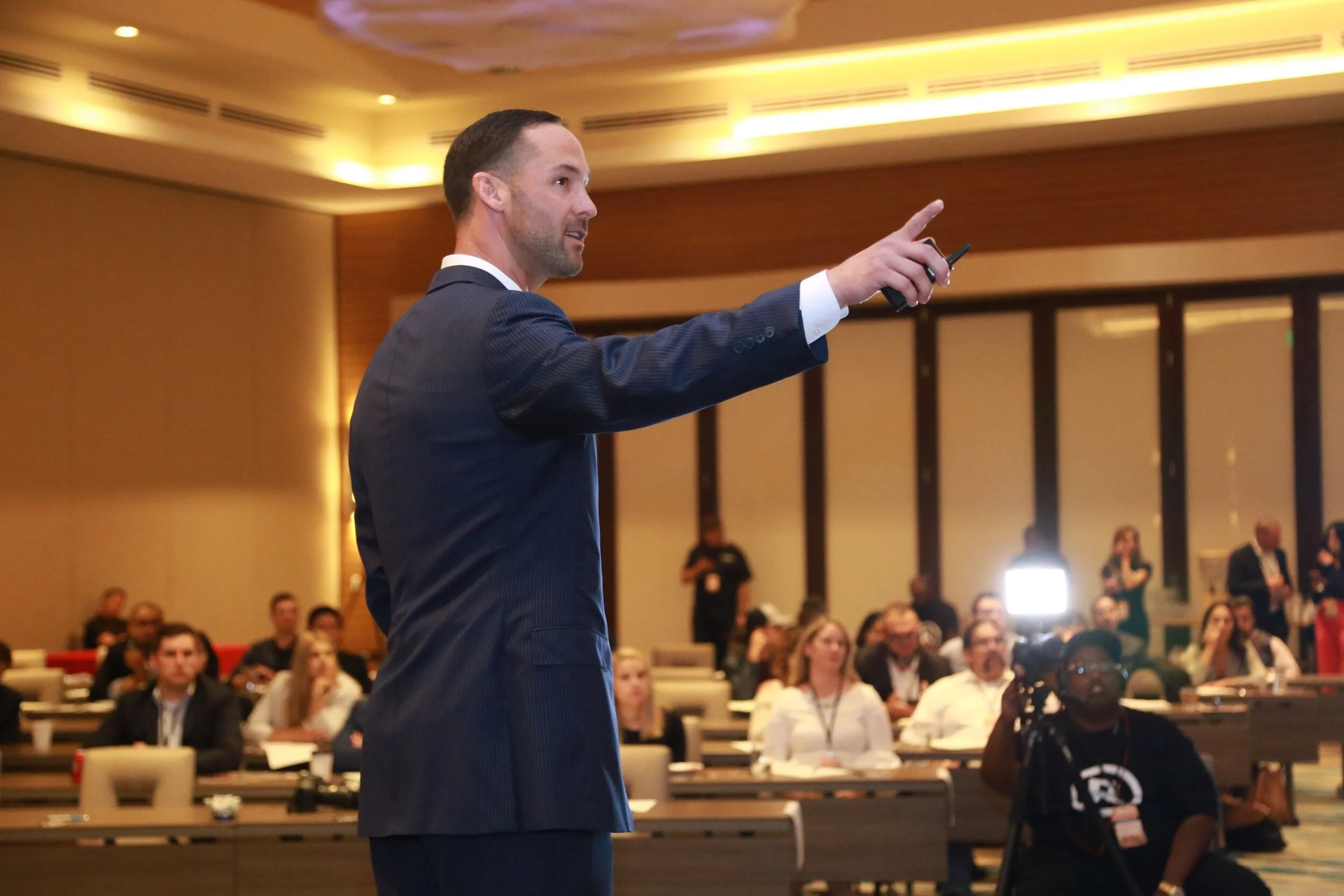 A man in a suit giving a presentation to an audience in a conference room. He is pointing with his hand, and there are people sitting at tables and listening attentively.
