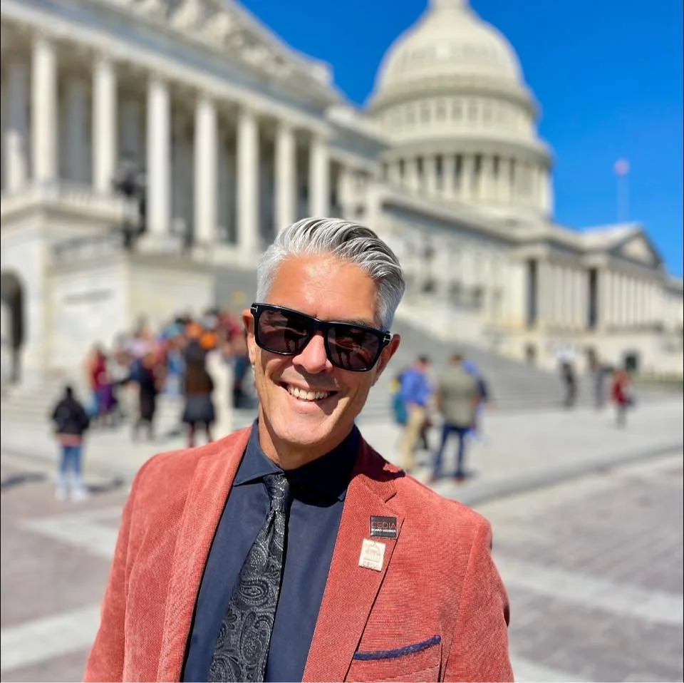 Smiling man with gray hair and sunglasses taking a selfie in front of the U.S. Capitol building with a blue sky and people in the background.