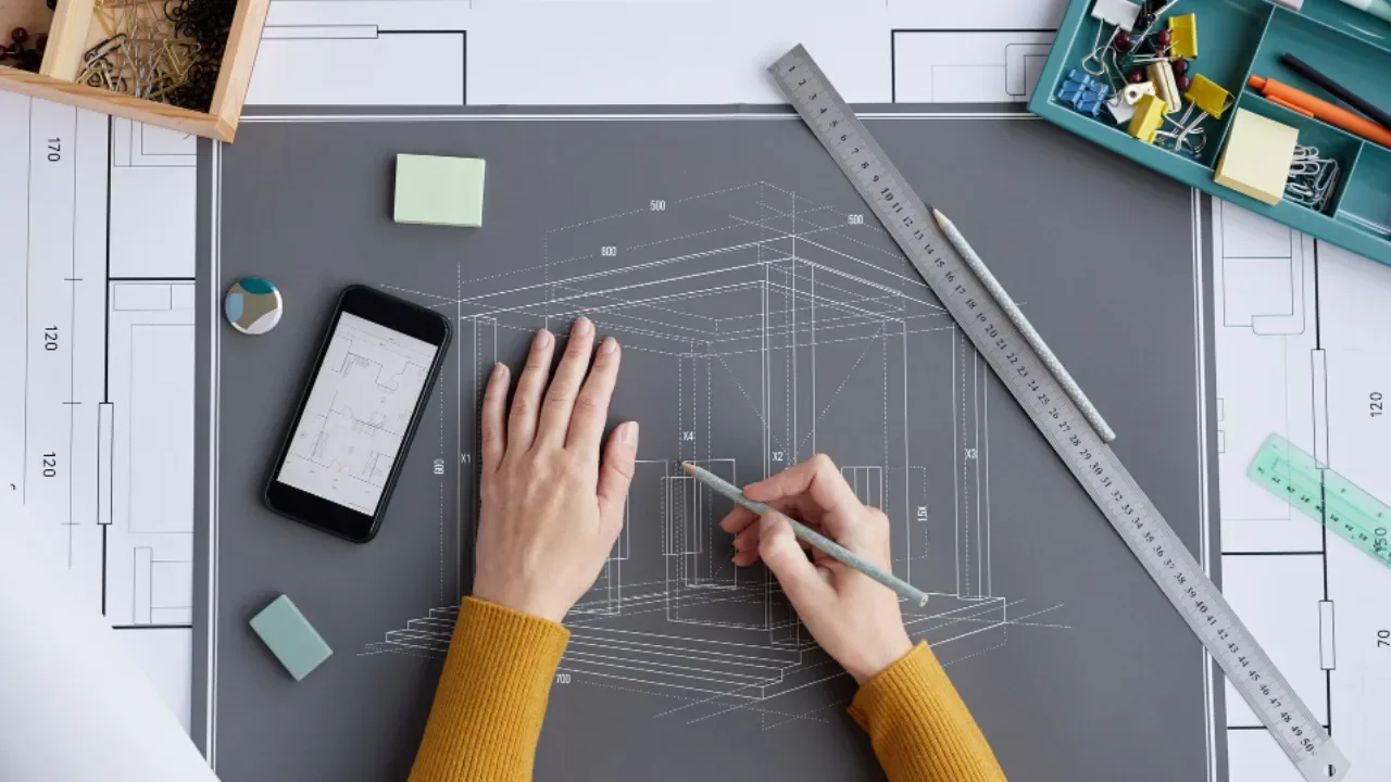 Architect working on a 3D house design on dark drafting paper with tools, a smartphone, and boxes of office supplies on the desk.