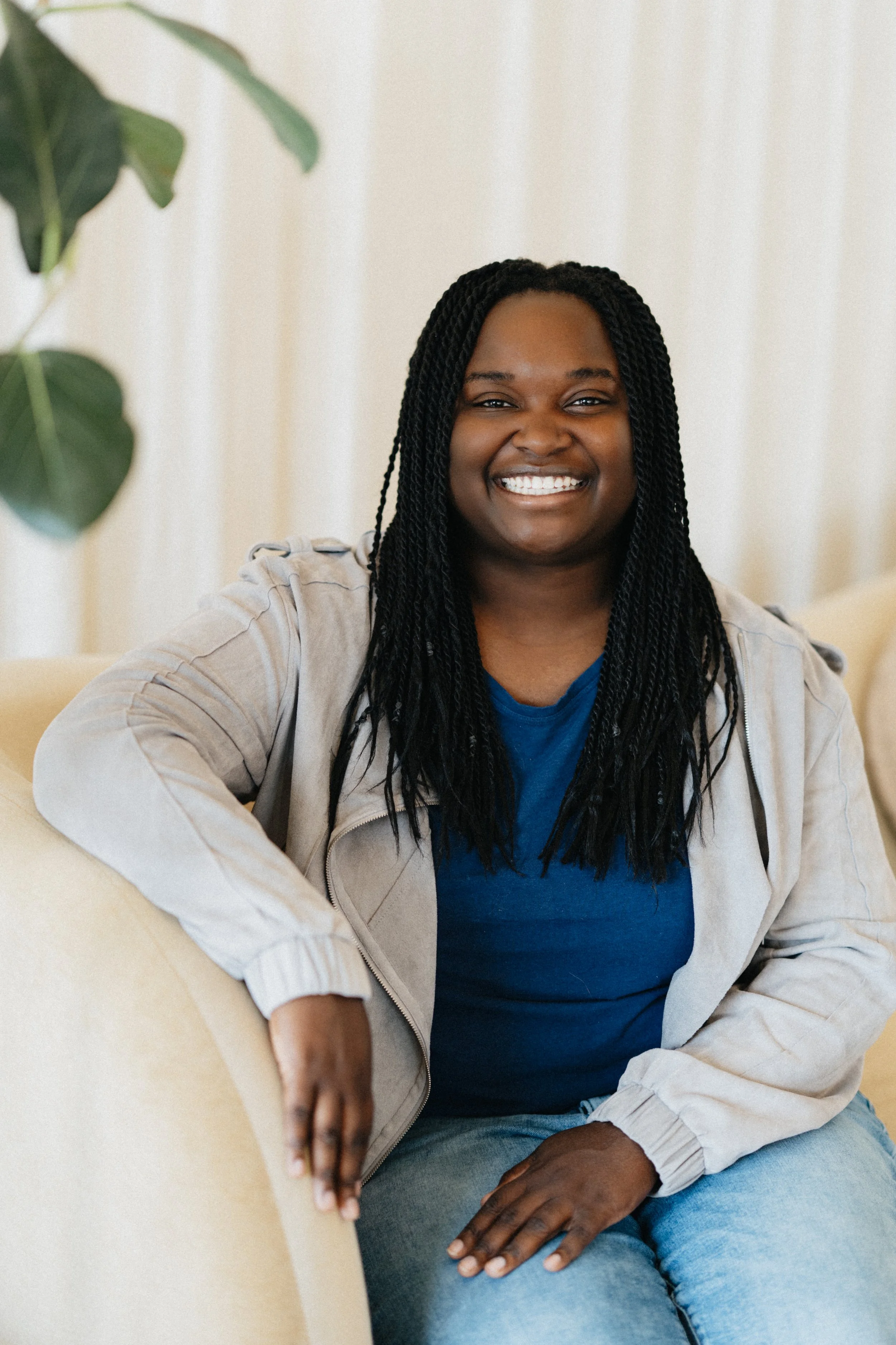 A smiling woman with long braided hair sitting on a black armchair in a room with white curtains and a potted plant.