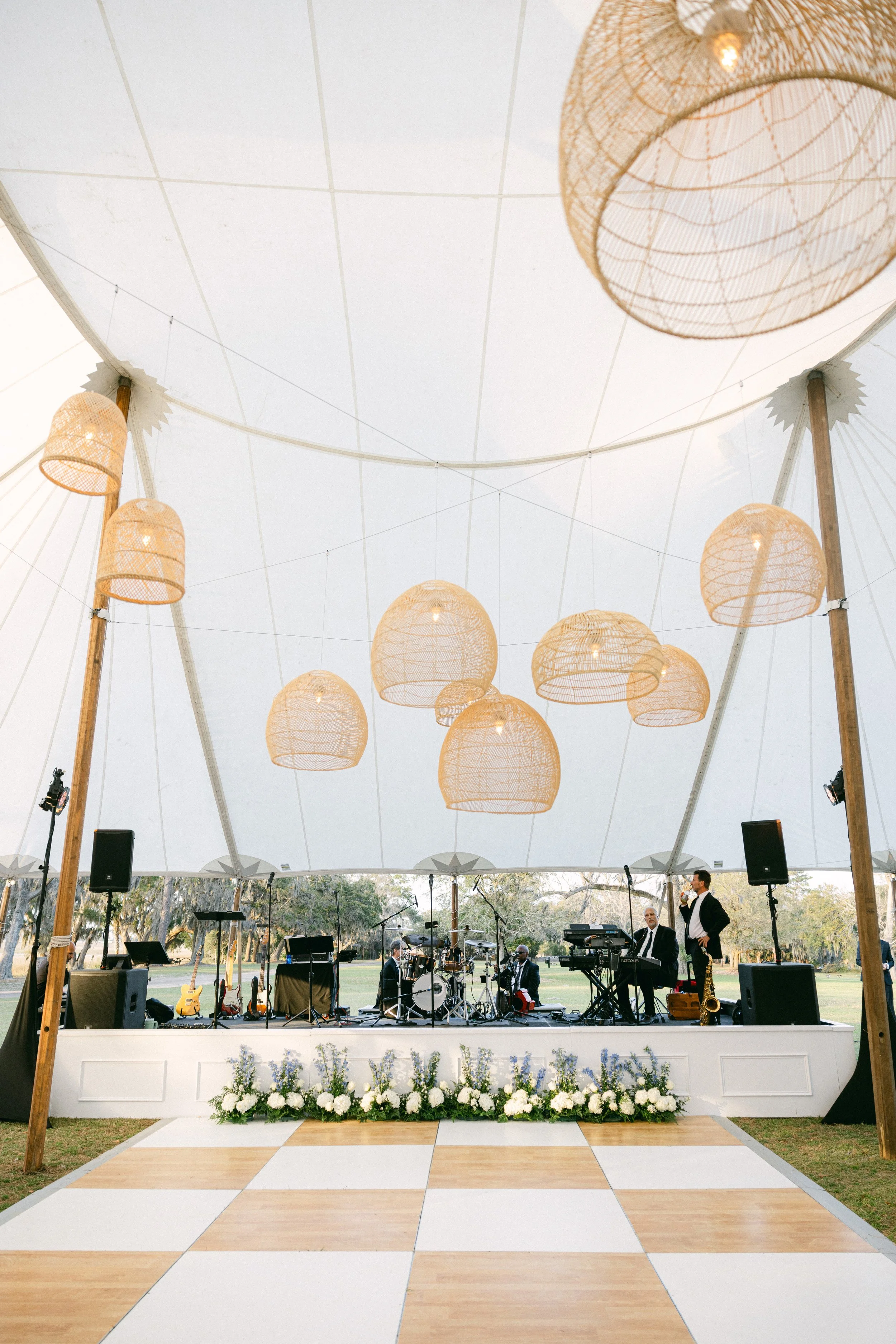 An outdoor stage set up under a large white tent with hanging woven lanterns, musical instruments, speakers, and a few people preparing for a performance.