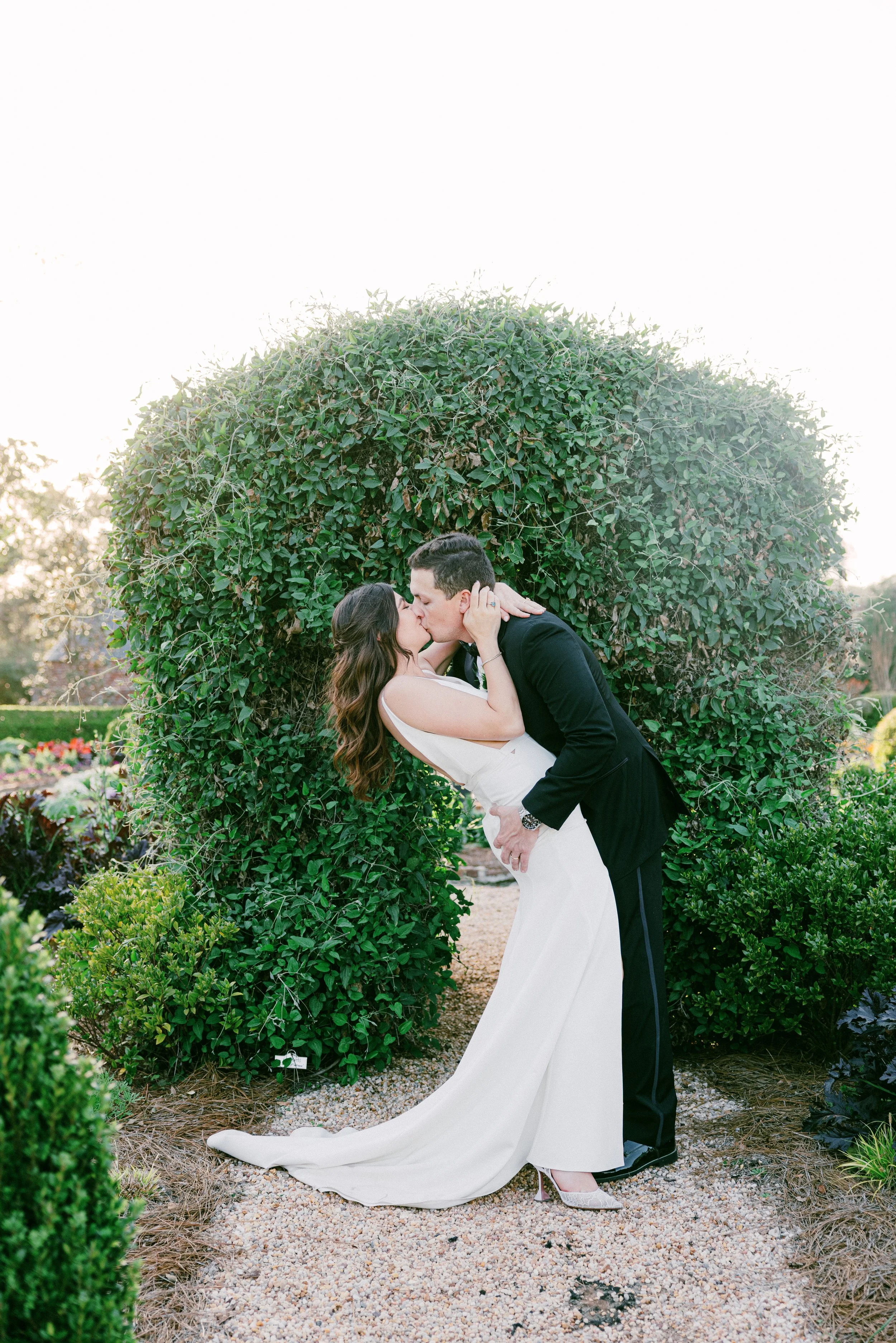 A bride and groom kissing outdoors beneath a large, rounded, green leafy shrub. The bride wears a white wedding dress and heels, the groom a black suit with a boutonniere. The scene is set in a garden with flowers and plants.
