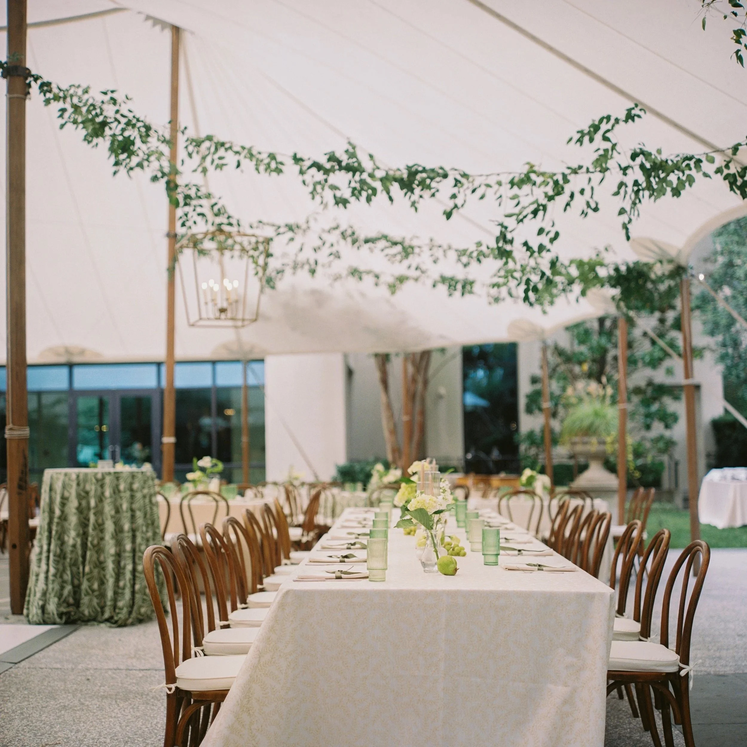 Long wedding reception table with textured linen, decorated with small flower arrangements and green apples, under a ceiling draped with greenery and a lantern.