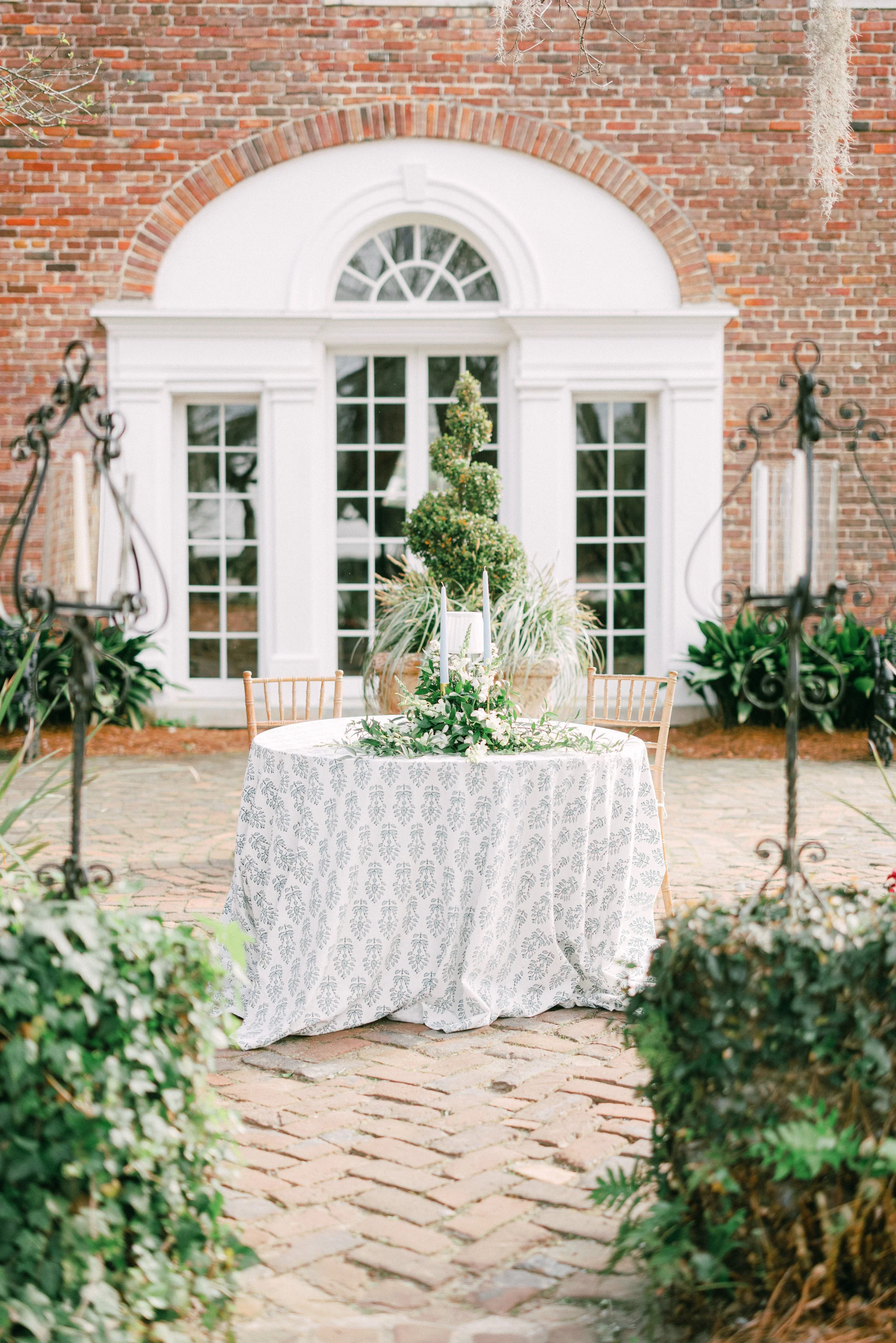 Outdoor wedding or special event setup with a round table covered with a patterned white tablecloth, decorated with greenery and white candles, placed on a cobblestone surface in front of a brick building with large white-framed windows and an arched