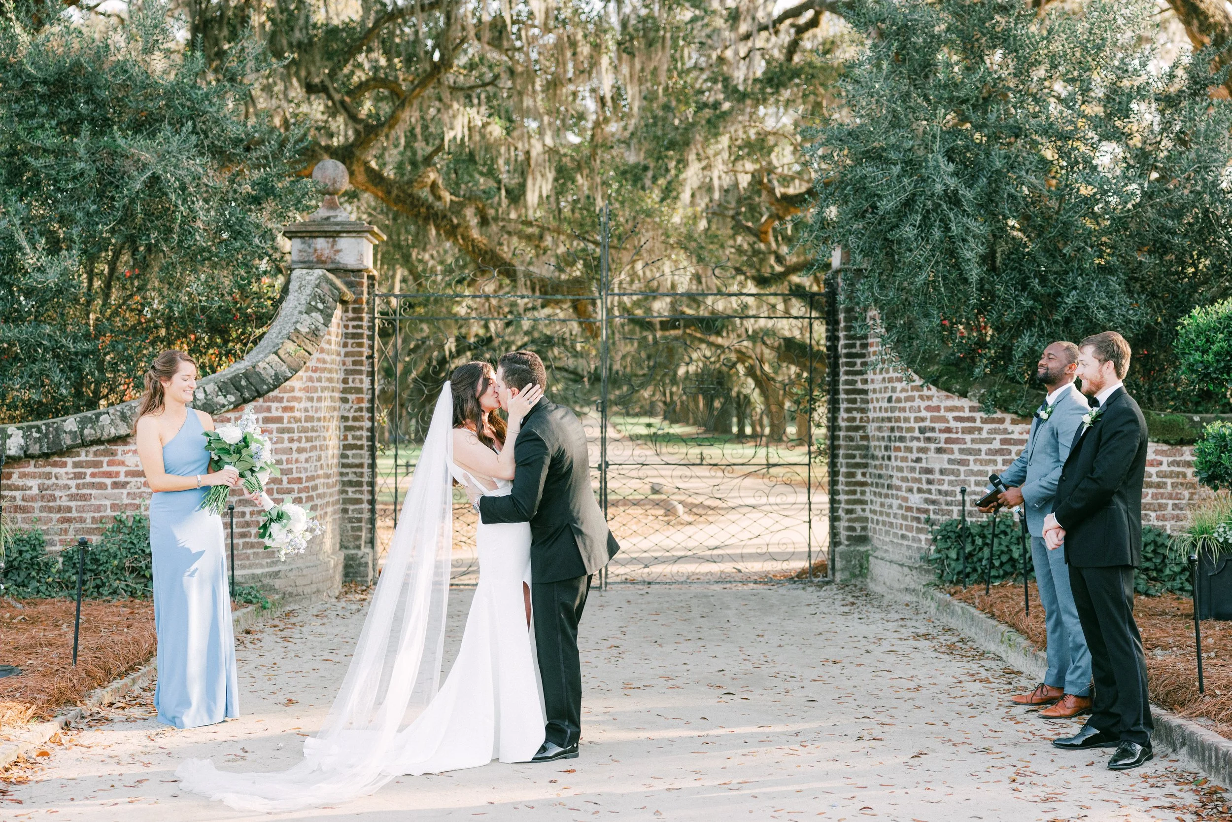 A wedding ceremony taking place outdoors with a bride and groom kissing, surrounded by wedding attendants and greenery.