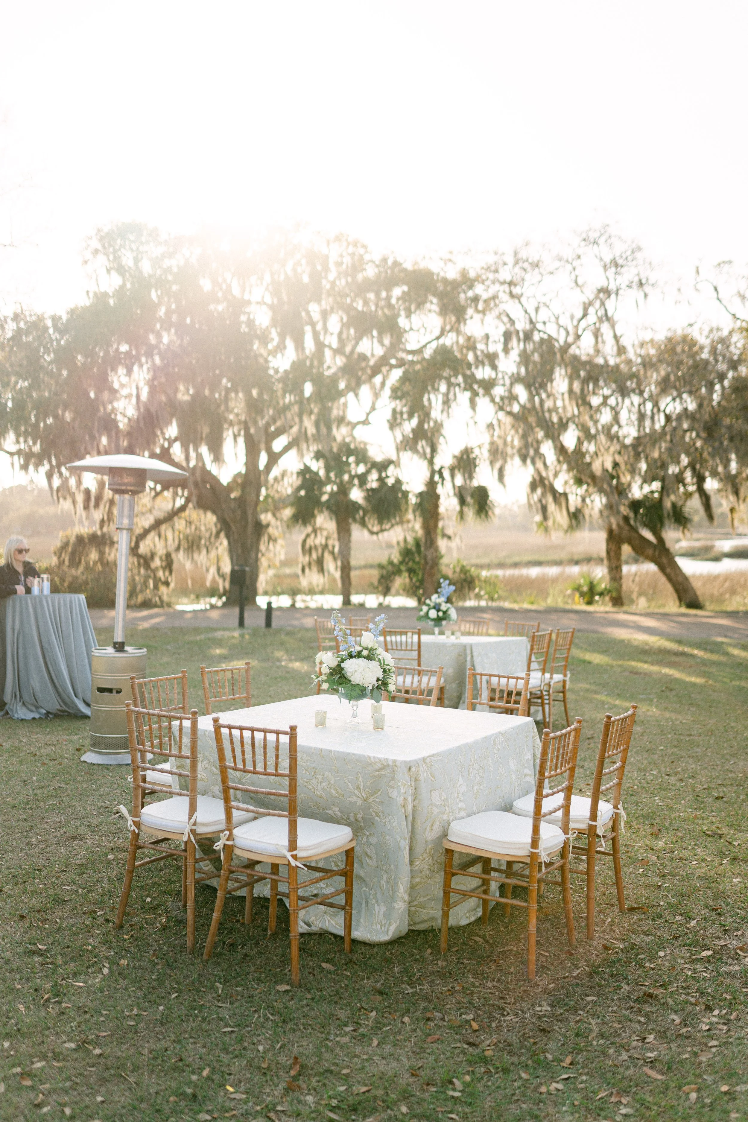 Outdoor wedding or event setup with a table adorned with flowers, surrounded by wooden chairs on grass, and trees in the background during sunset.