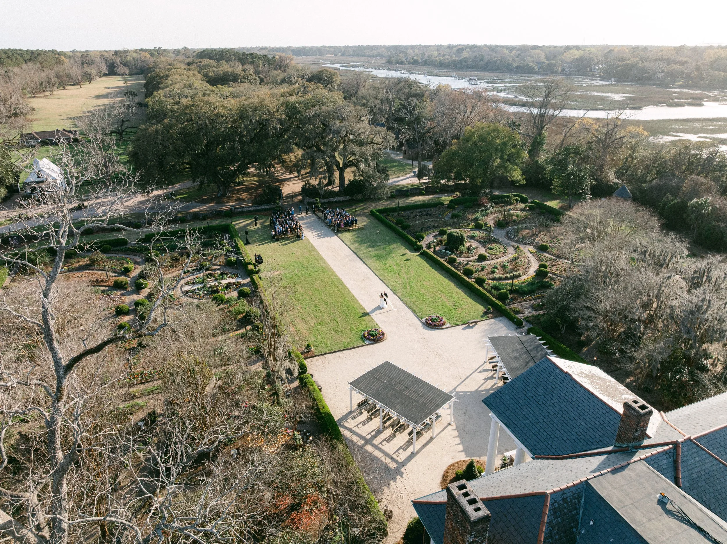 An aerial view of a wedding ceremony outdoors on a lawn with trees and a garden, near a river or wetland area.