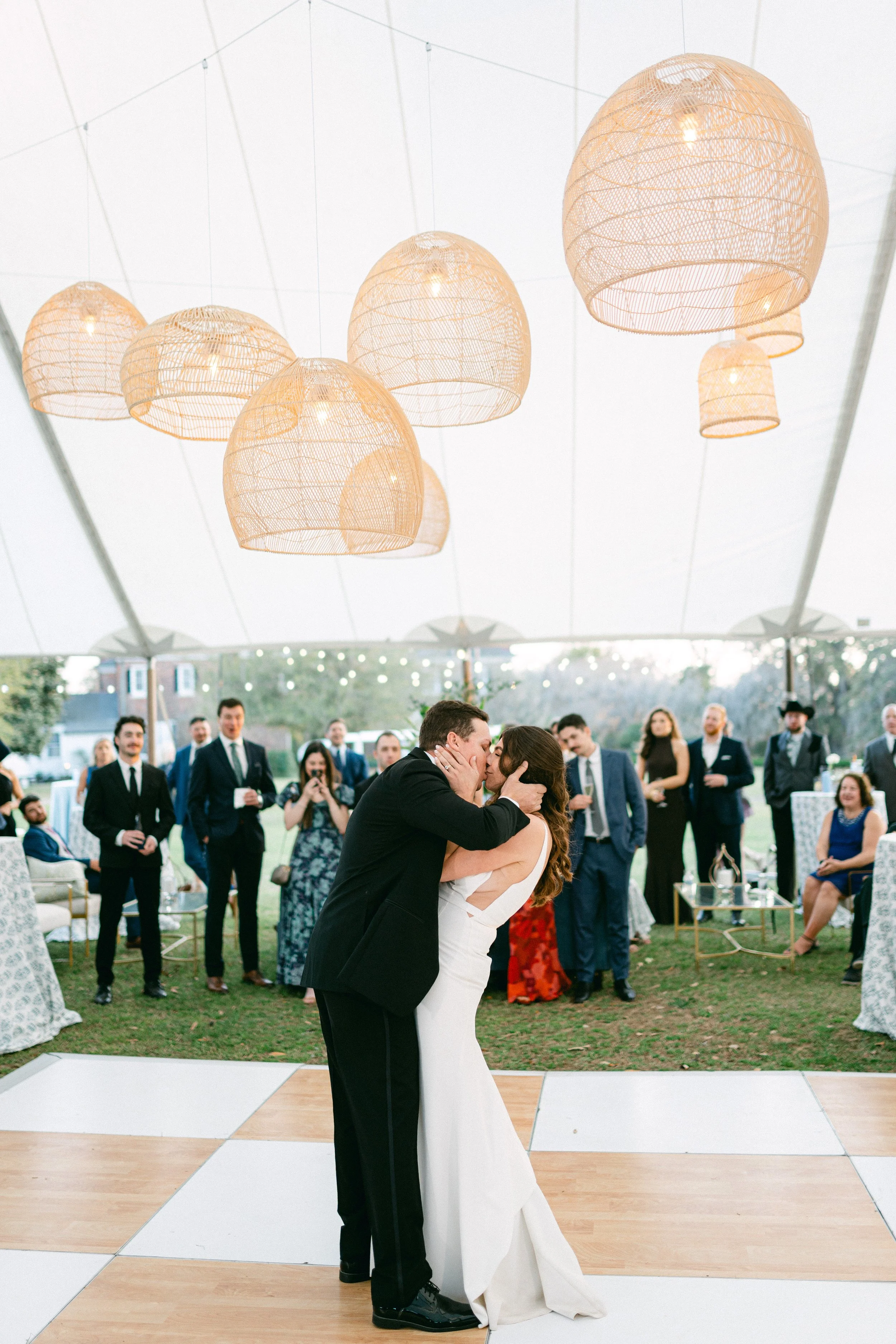 A bride and groom dance together at their wedding reception, surrounded by guests under a tent with hanging woven light fixtures.
