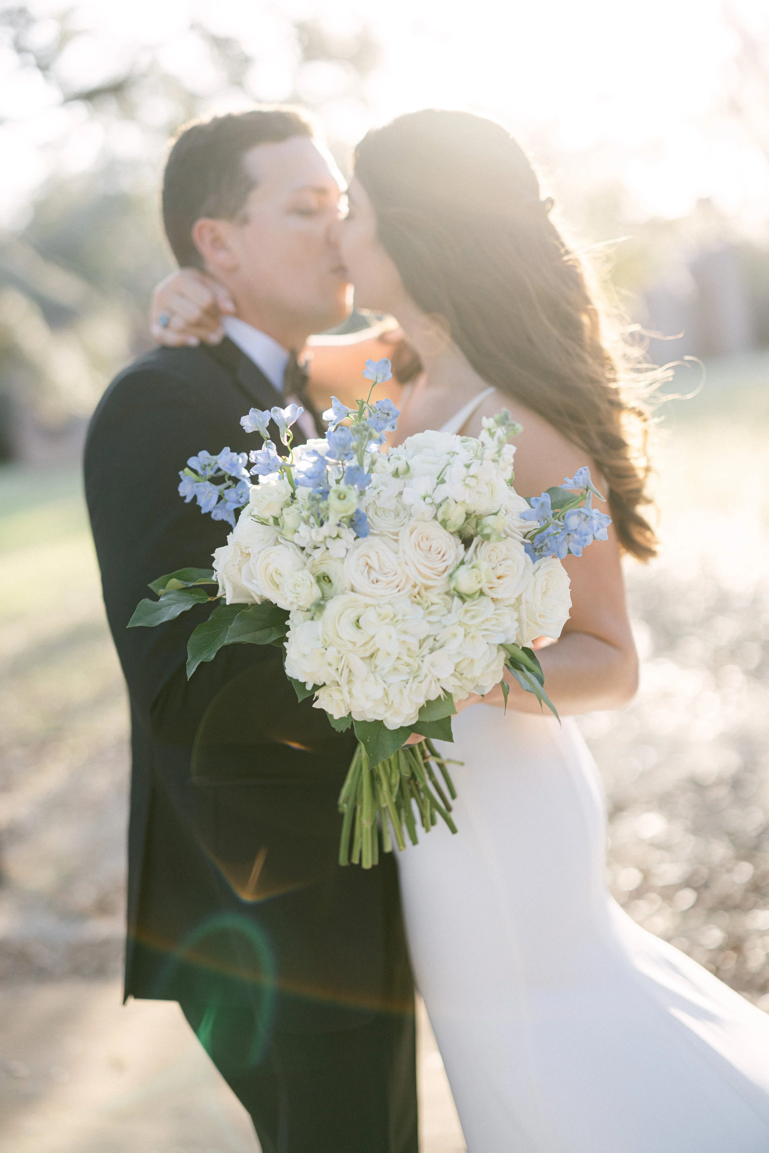 A gay couple in formal wedding attire sharing a kiss outdoors during sunlight, with one holding a bouquet of white and blue flowers.