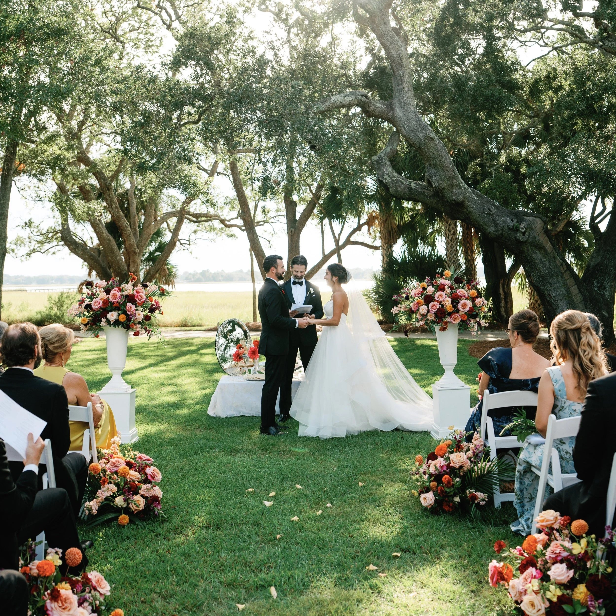 A wedding ceremony outdoors under large trees, with a bride and groom holding hands, surrounded by bright floral arrangements and seated guests.