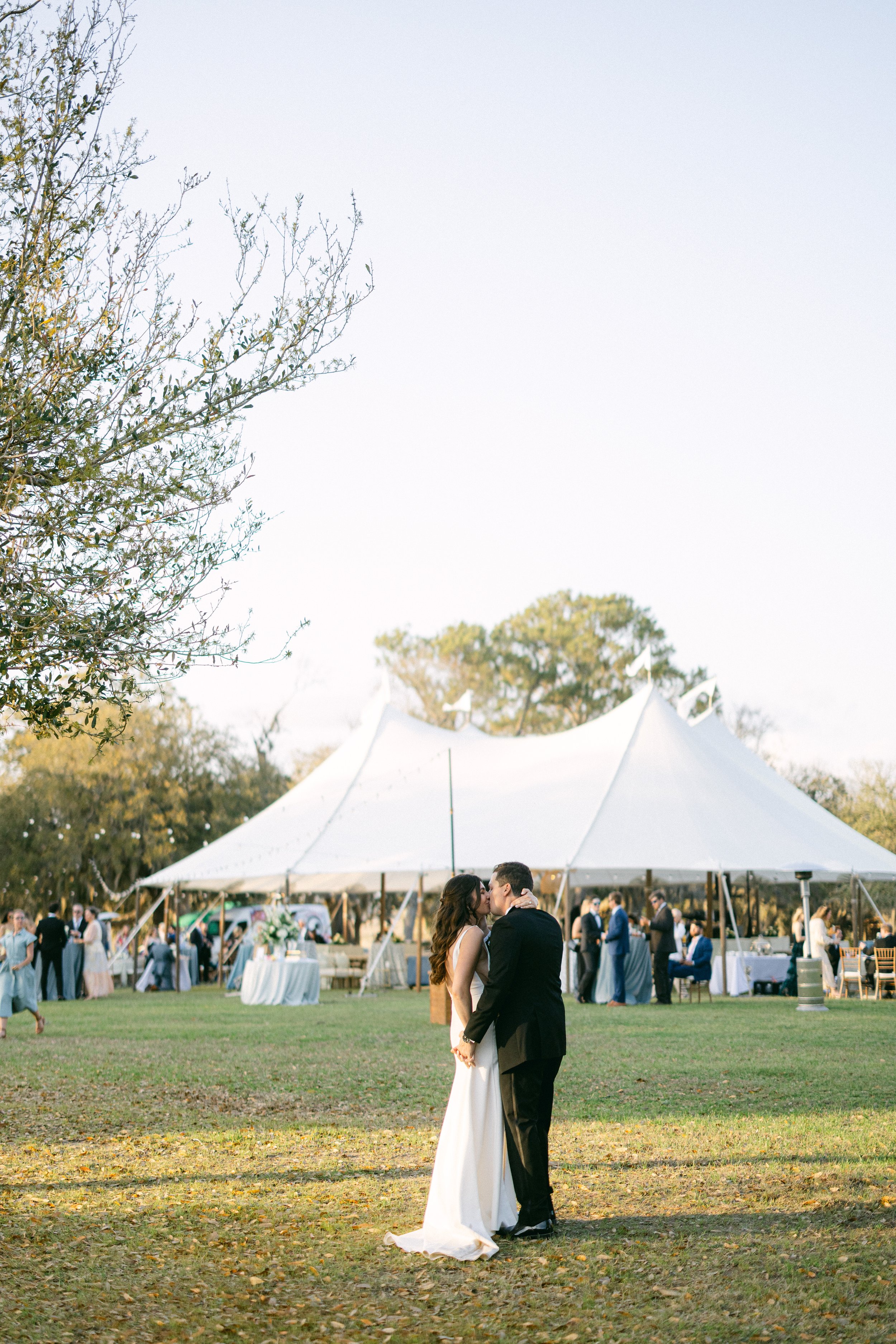 A bride and groom sharing a kiss at an outdoor wedding ceremony with a white canopy, guests in the background, and trees on a sunny day.