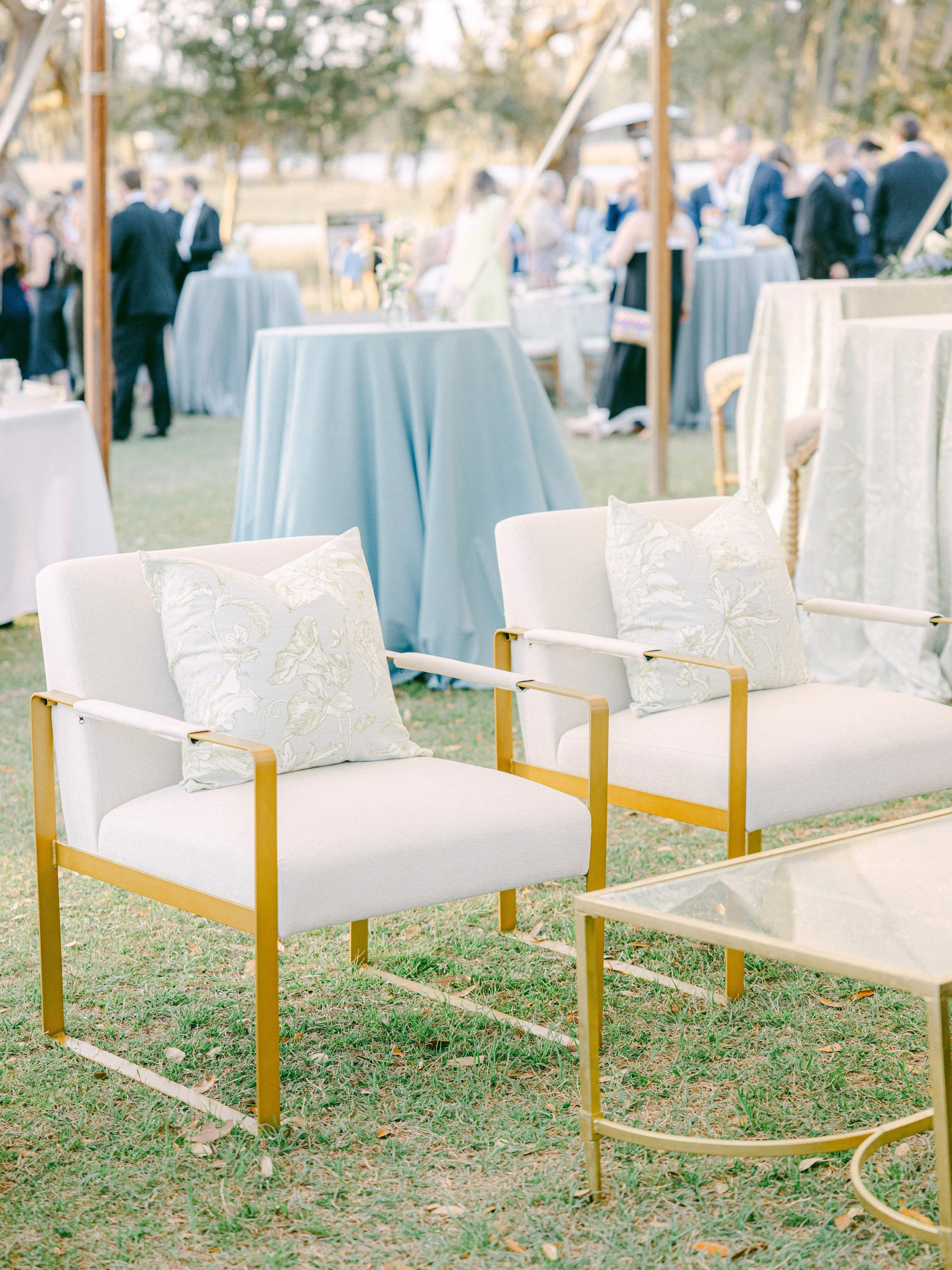 Outdoor wedding reception area with white and gold chairs, some with floral pillows, on grass with tall tables draped in pastel blue tablecloths, and a group of people in formal attire in the background under a wooden frame.