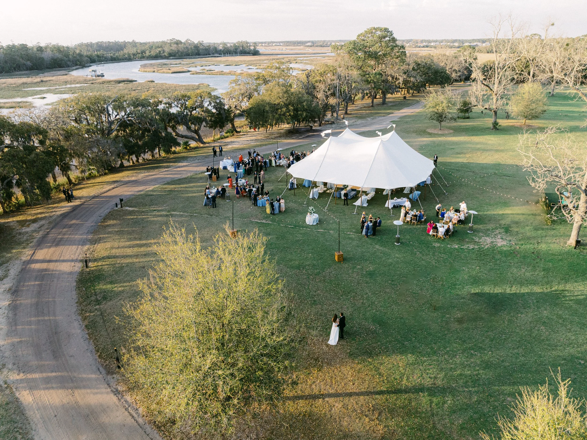 An outdoor wedding reception under a large white tent in a scenic, grassy area by a river with trees and wetlands in the background. Guests are mingling and seated at tables, with some standing near the tent.