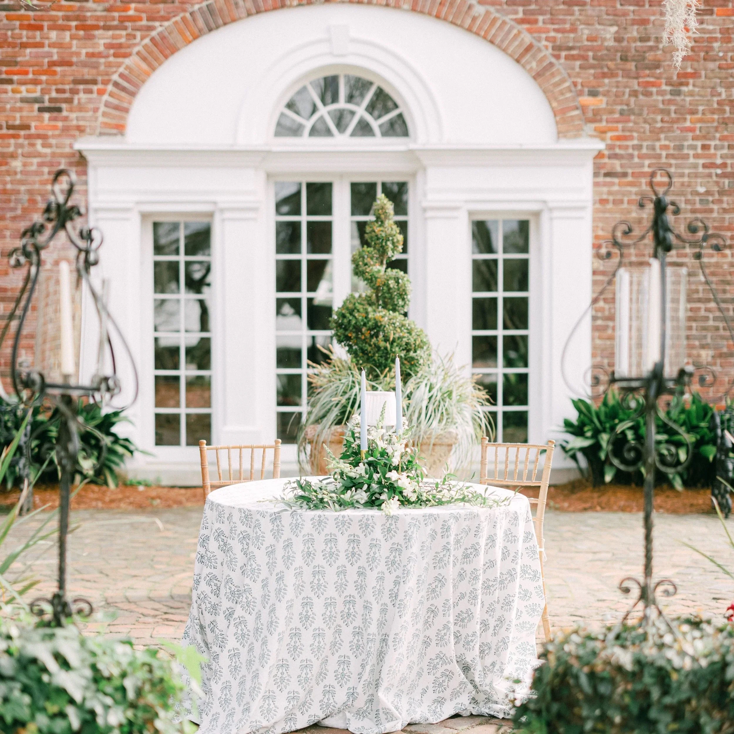 A white table covered with a white tablecloth with a gray floral pattern. The table is set outdoors with green plants surrounding it. In the background, there is a large white window with multiple panes, framed by a brick exterior wall. A small, lush topiary plant is centered on the table.