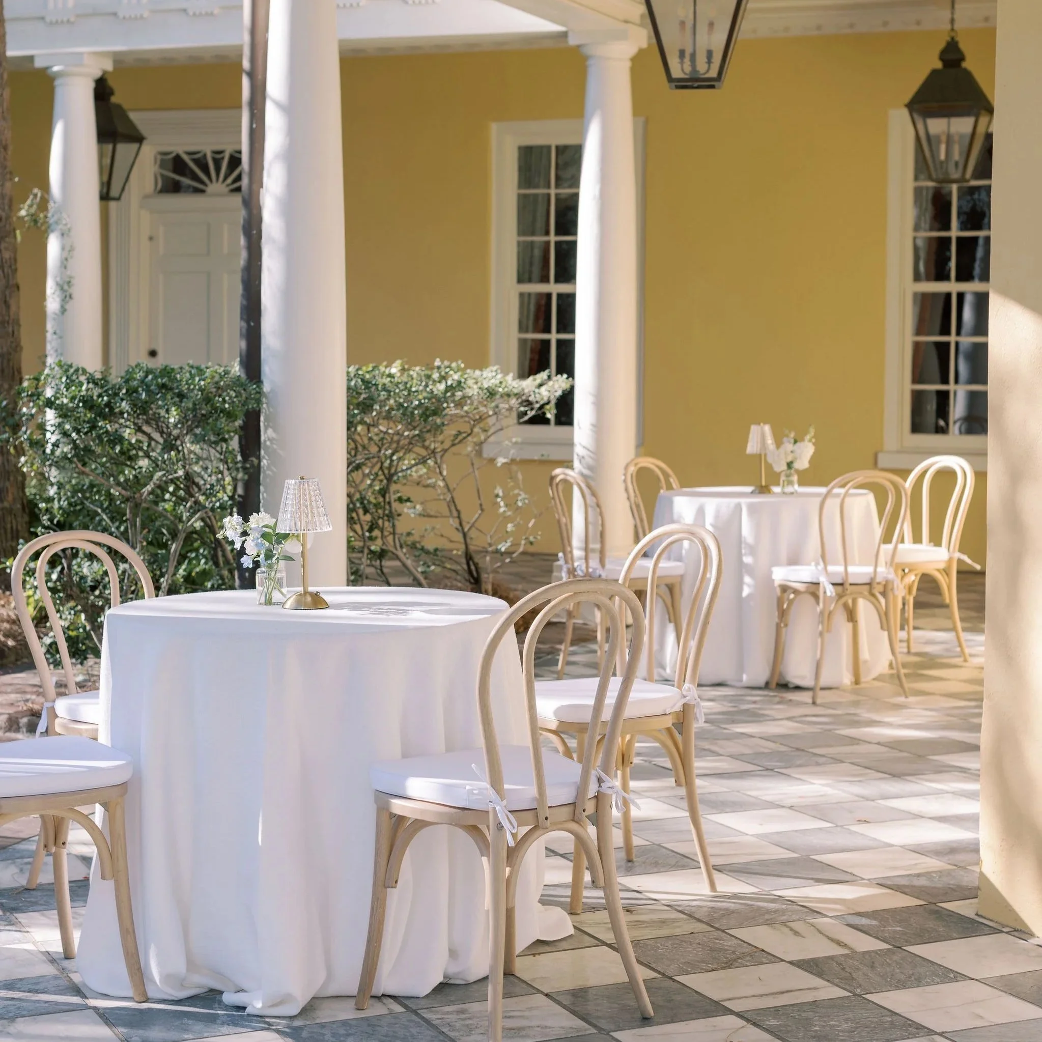 Outdoor dining area with round tables covered with white linens, surrounded by white washed chairs, with small lamps and floral centerpieces. The setting is the William Aiken House with their lanterns hanging overhead.