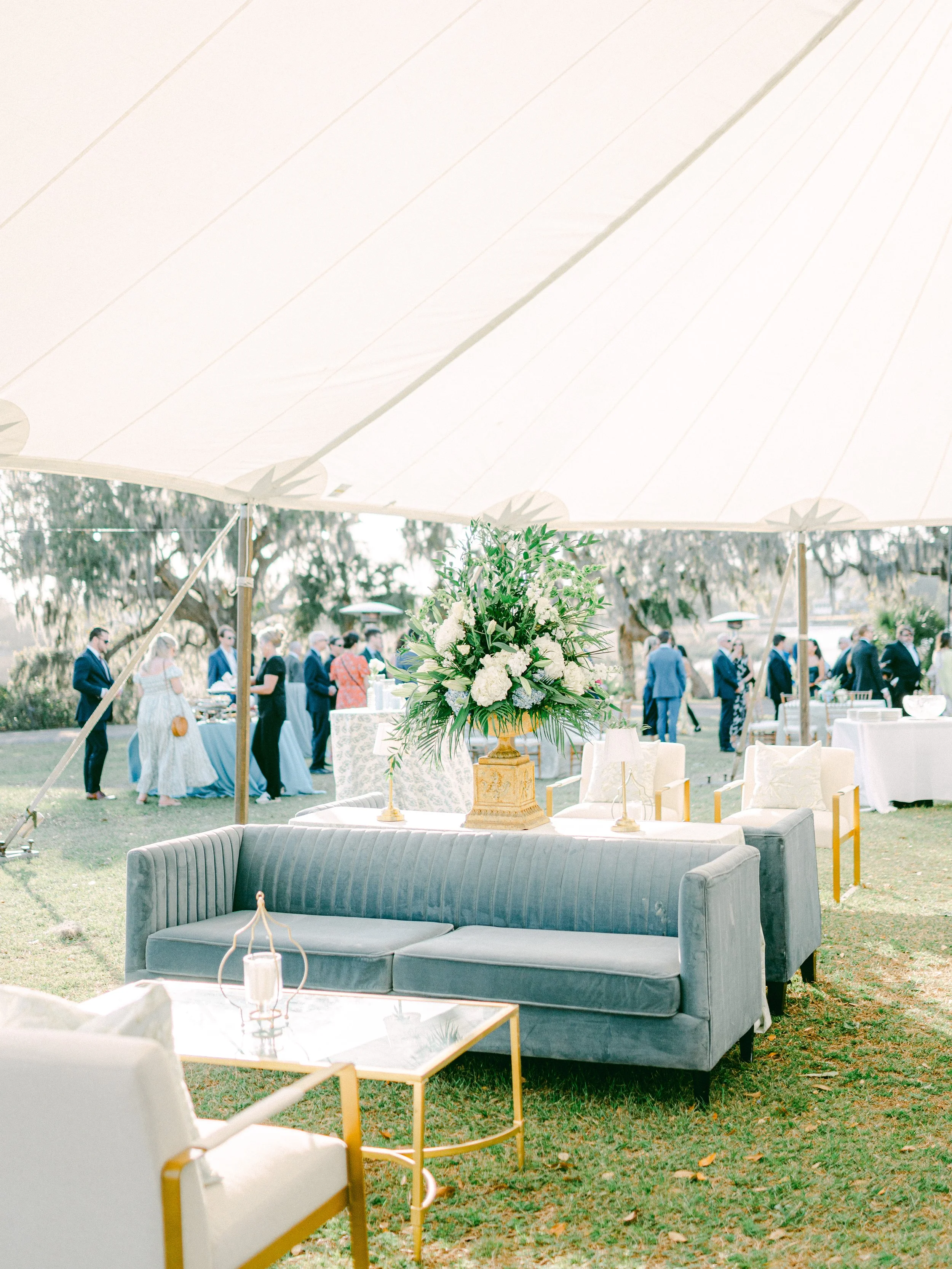 An outdoor event setup under a large white canopy tent with a floral centerpiece on a yellow pedestal, surrounded by white and gray seating, with people mingling in the background on a grassy area.