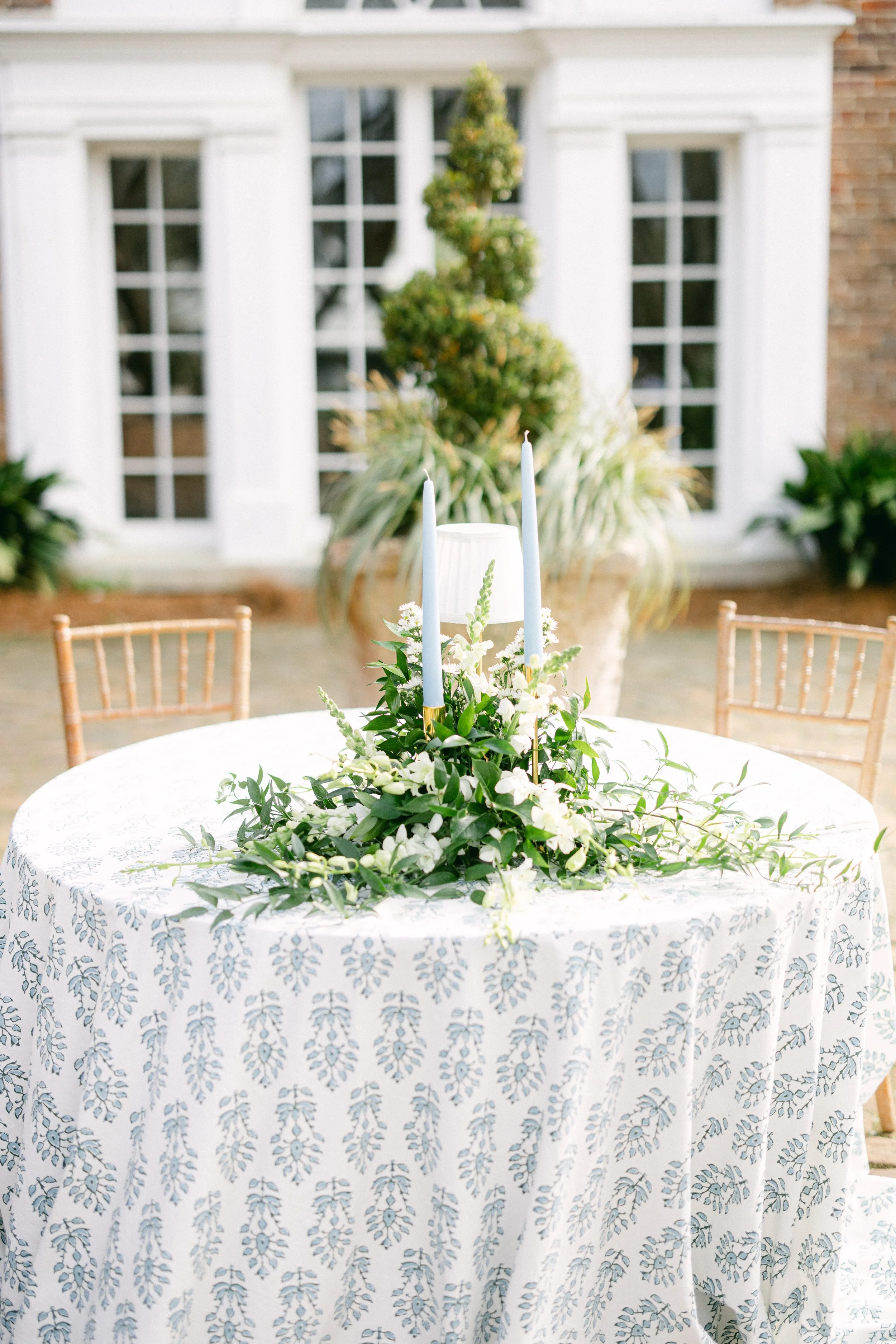 Outdoor event table decorated with white flowers and blue candles, surrounded by gold chairs, with a background of a building with white walls and windows.