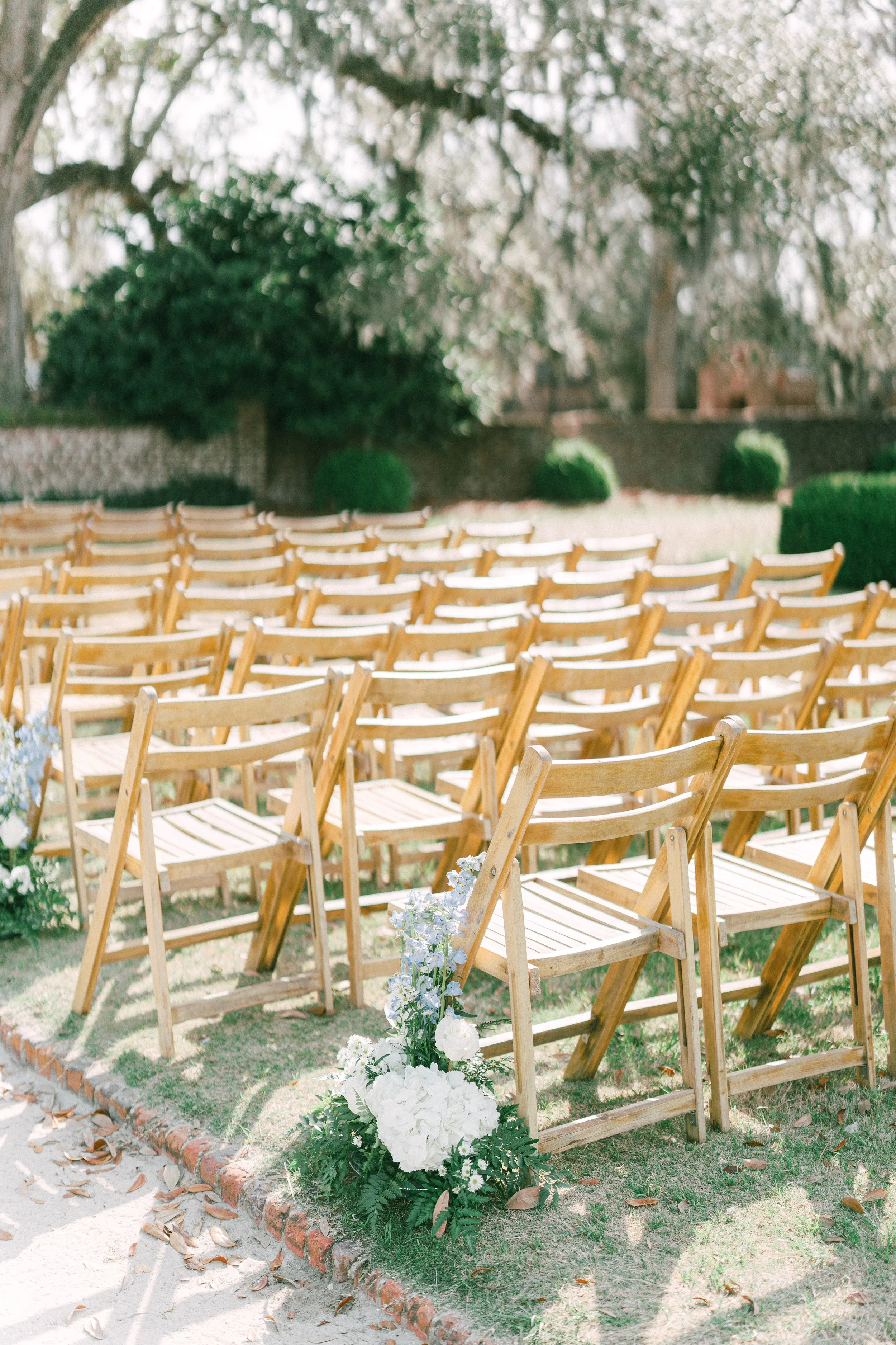 Outdoor wedding ceremony setup with multiple wooden folding chairs arranged in rows, decorated with white flowers at the front, and surrounded by trees and bushes in a garden setting.