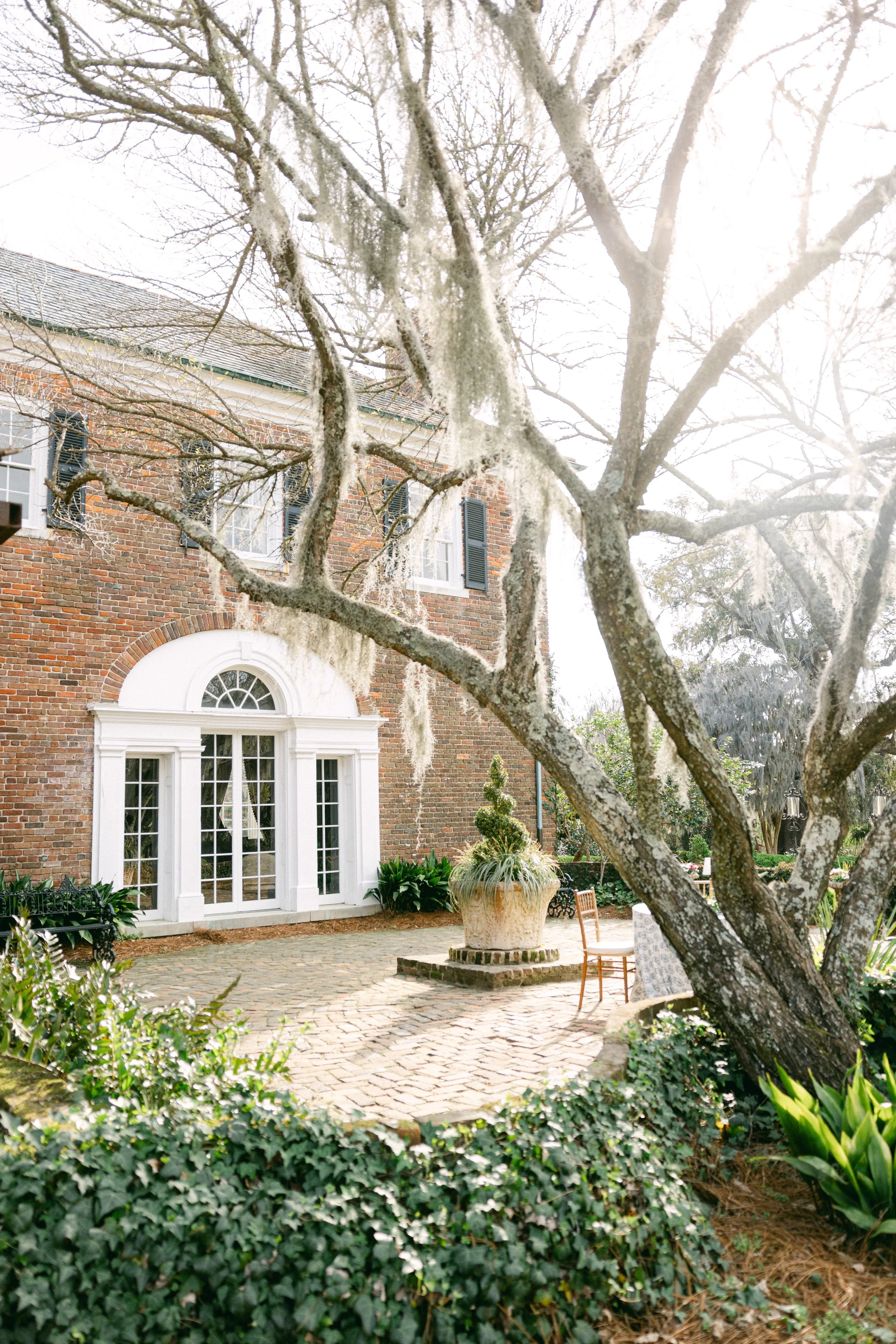 A brick house with white framed glass doors, black shutters, and a row of windows, with a tree in the foreground and a garden patio with chairs, a large planter, and lush greenery.