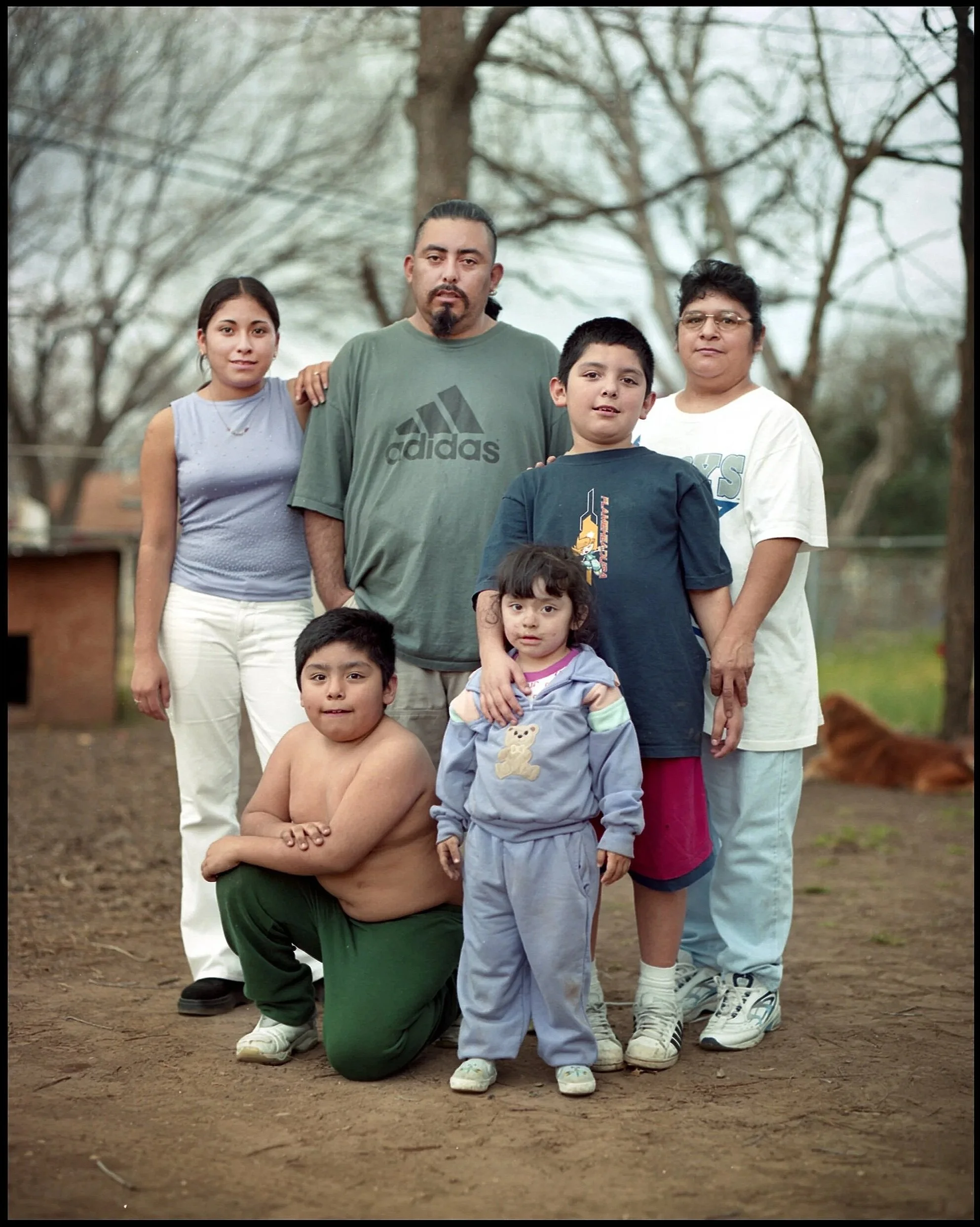 Group photo of a diverse family of seven people outdoors in a park, standing on dirt ground with leafless trees in the background, some adults and children smiling or with neutral expressions.