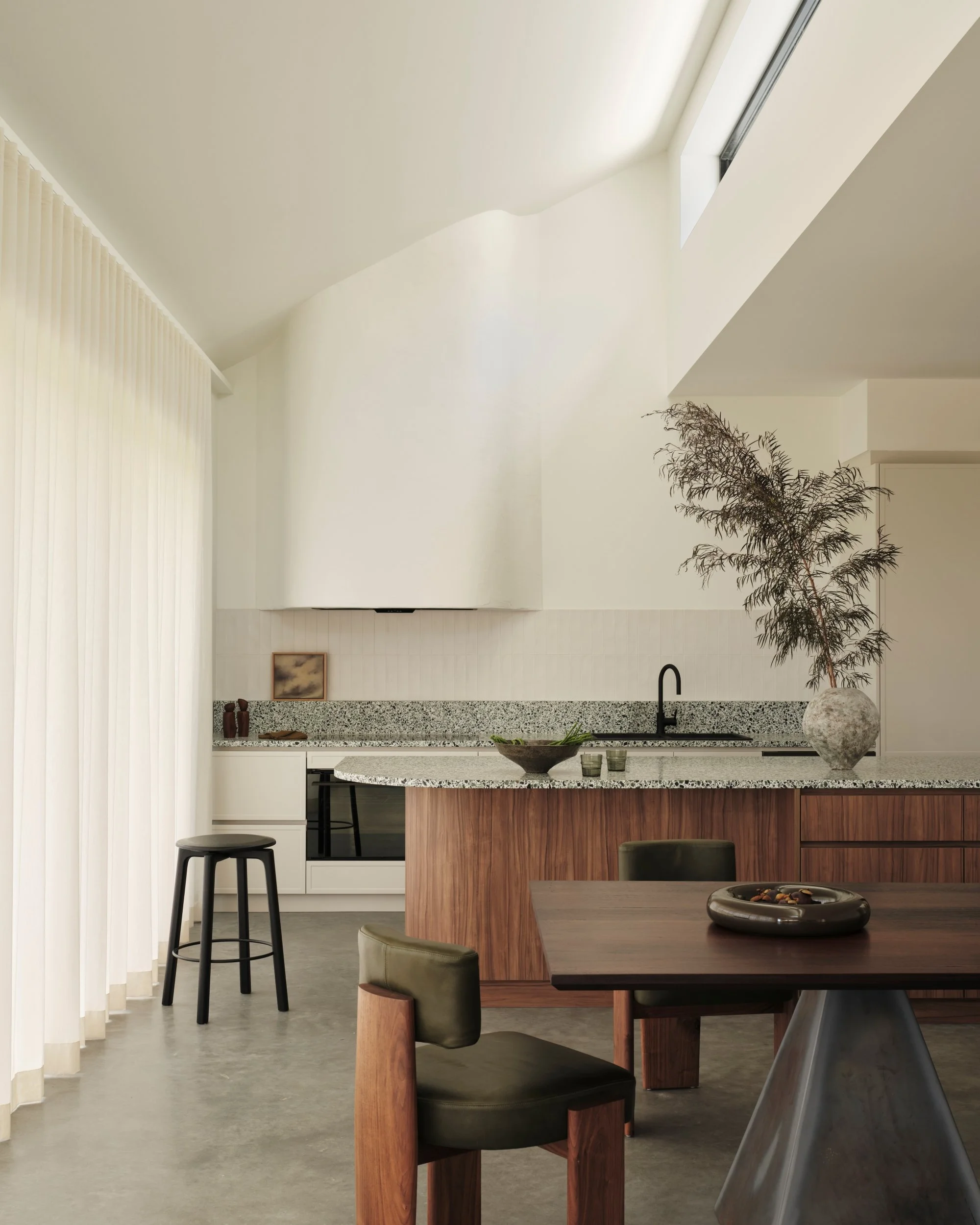 Modern coastal kitchen with terrazzo benchtops, curved plaster range hood, timber island cabinetry and minimalist black tapware, styled with natural decor and soft daylight through sheer curtains.