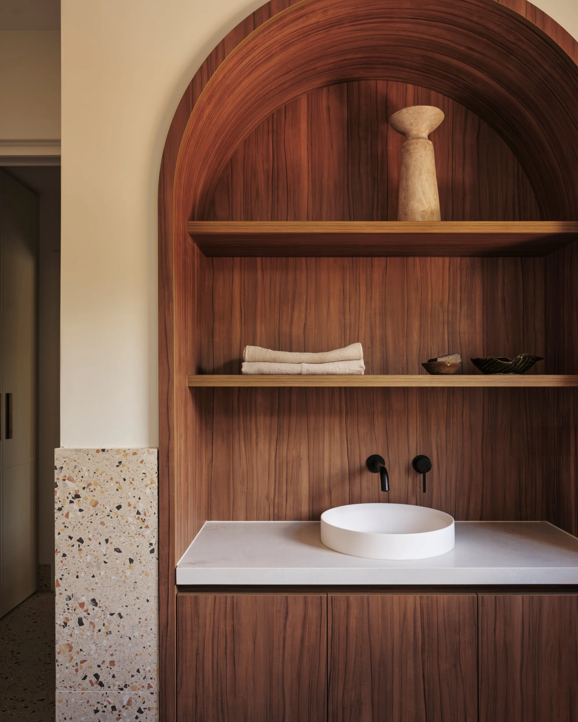 Custom timber vanity with arched shelving niche, round basin and wall-mounted black tapware, paired with terrazzo detail and warm walnut joinery in a modern Australian bathroom design.