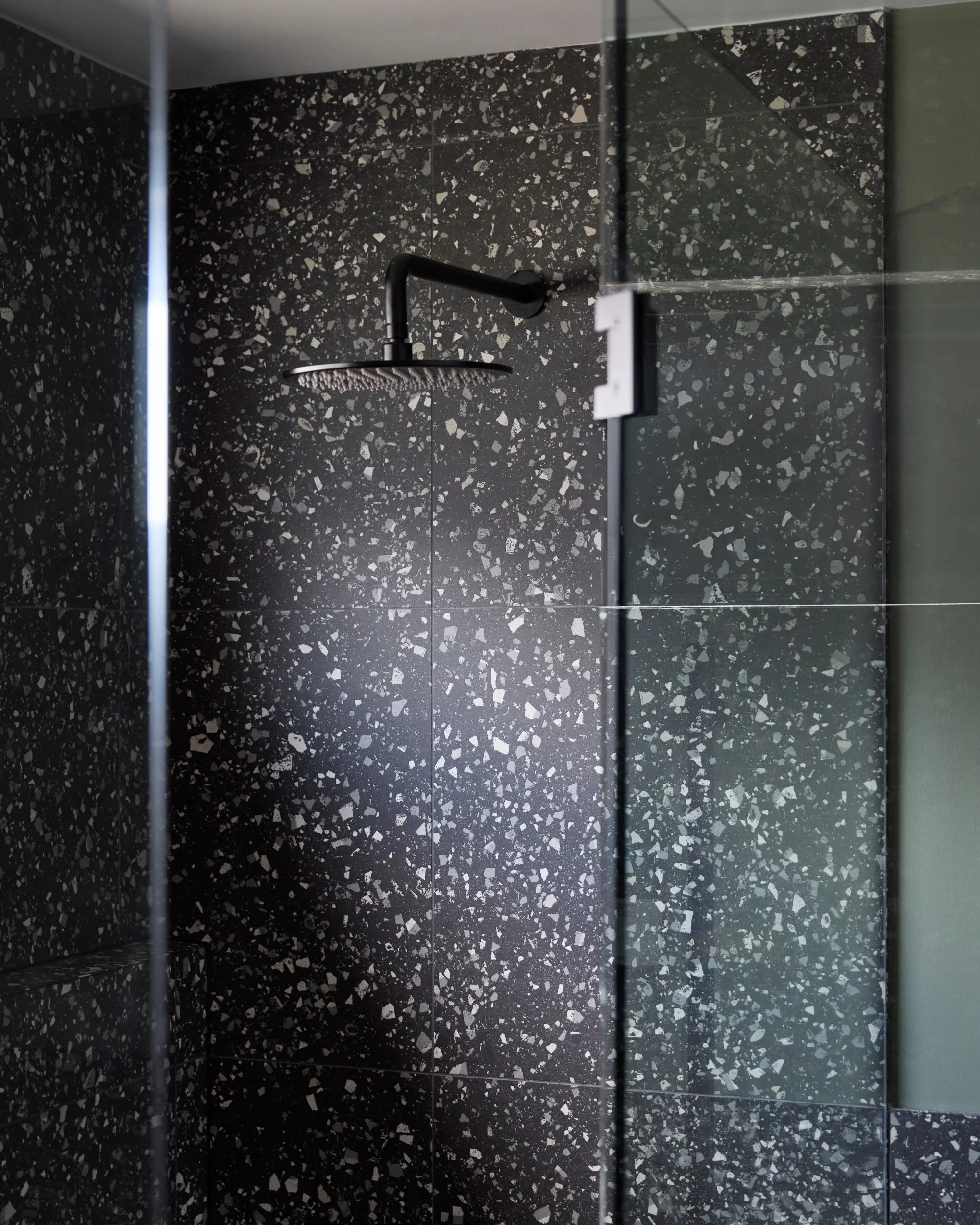 Modern bathroom shower with terrazzo walls and matte black rain shower fixture behind a frameless glass screen, highlighting texture and contrast in contemporary design.