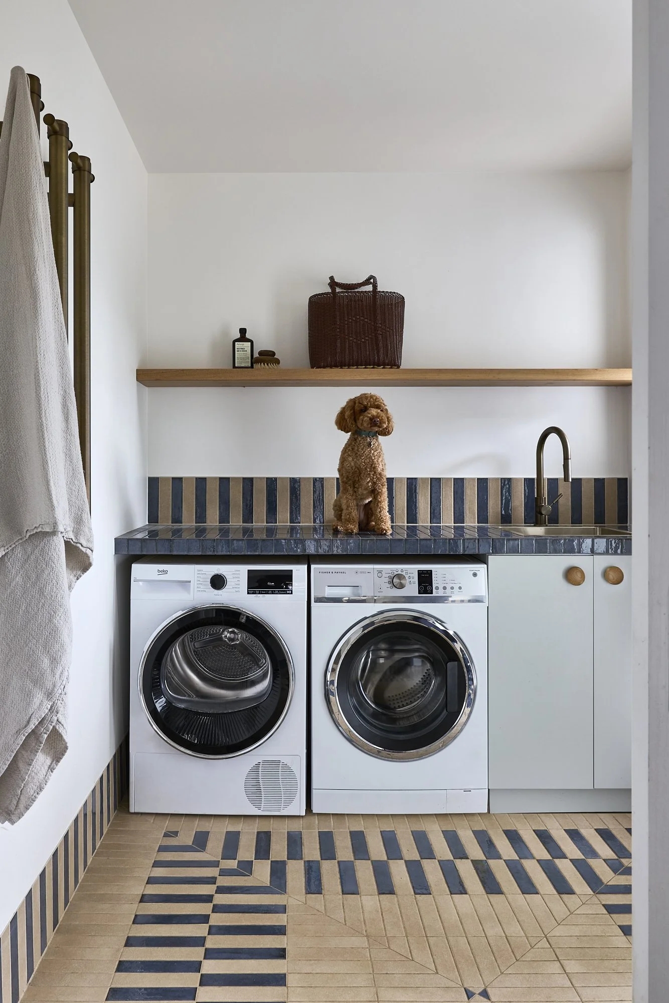 Contemporary Australian laundry featuring patterned blue and beige floor tiles, timber floating shelf, white cabinetry, integrated sink, brass tapware and custom joinery in a coastal interior by Revel Design.