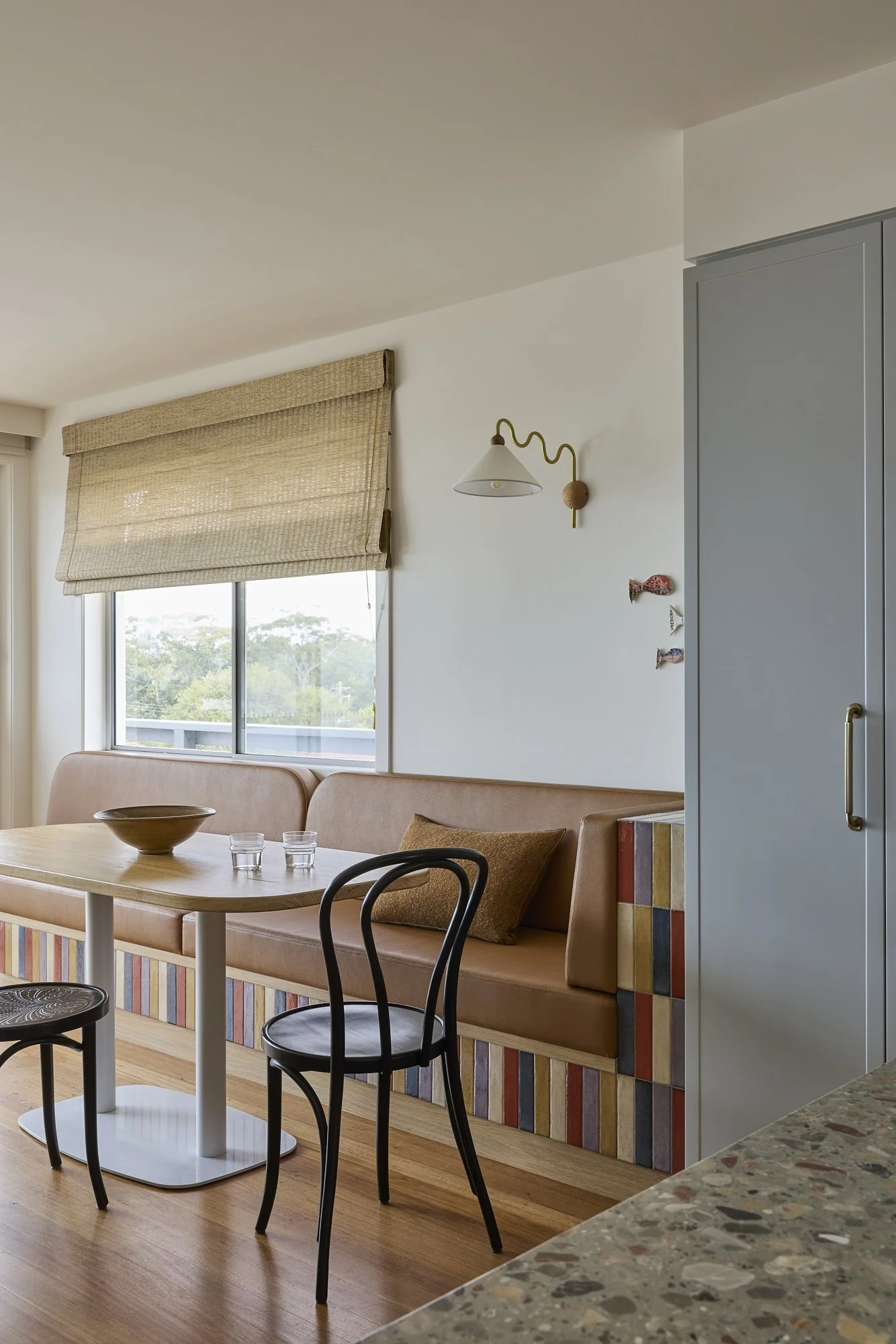 Contemporary dining nook featuring custom leather banquette seating, handcrafted colourful tiled joinery, timber table and woven Roman blind in an Australian family home.