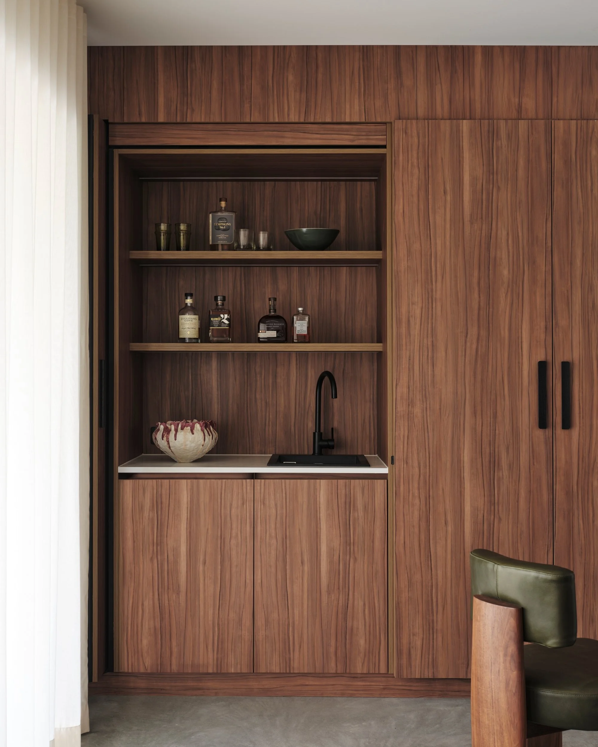 Custom walnut bar cabinet with open shelving, integrated sink and matte black tapware, styled with glassware and bottles in a refined modern interior joinery detail.