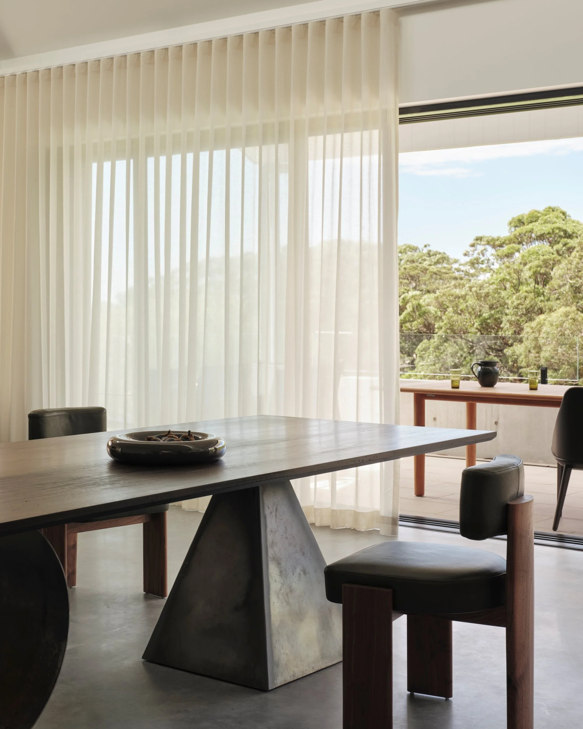Modern dining room with sculptural timber table, leather dining chairs and floor-to-ceiling sheer curtains opening to a balcony with treetop views, blending indoor and outdoor coastal living.